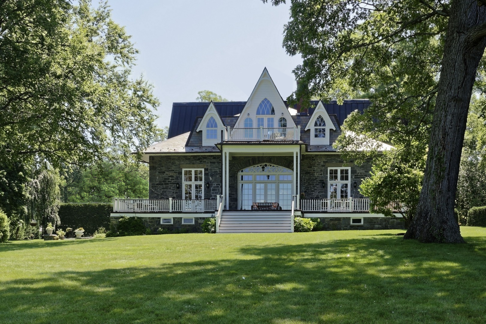 A large, historic, American Gothic stone house with white trim, multiple gabled windows, and a central balcony, surrounded by lush green trees and grass.