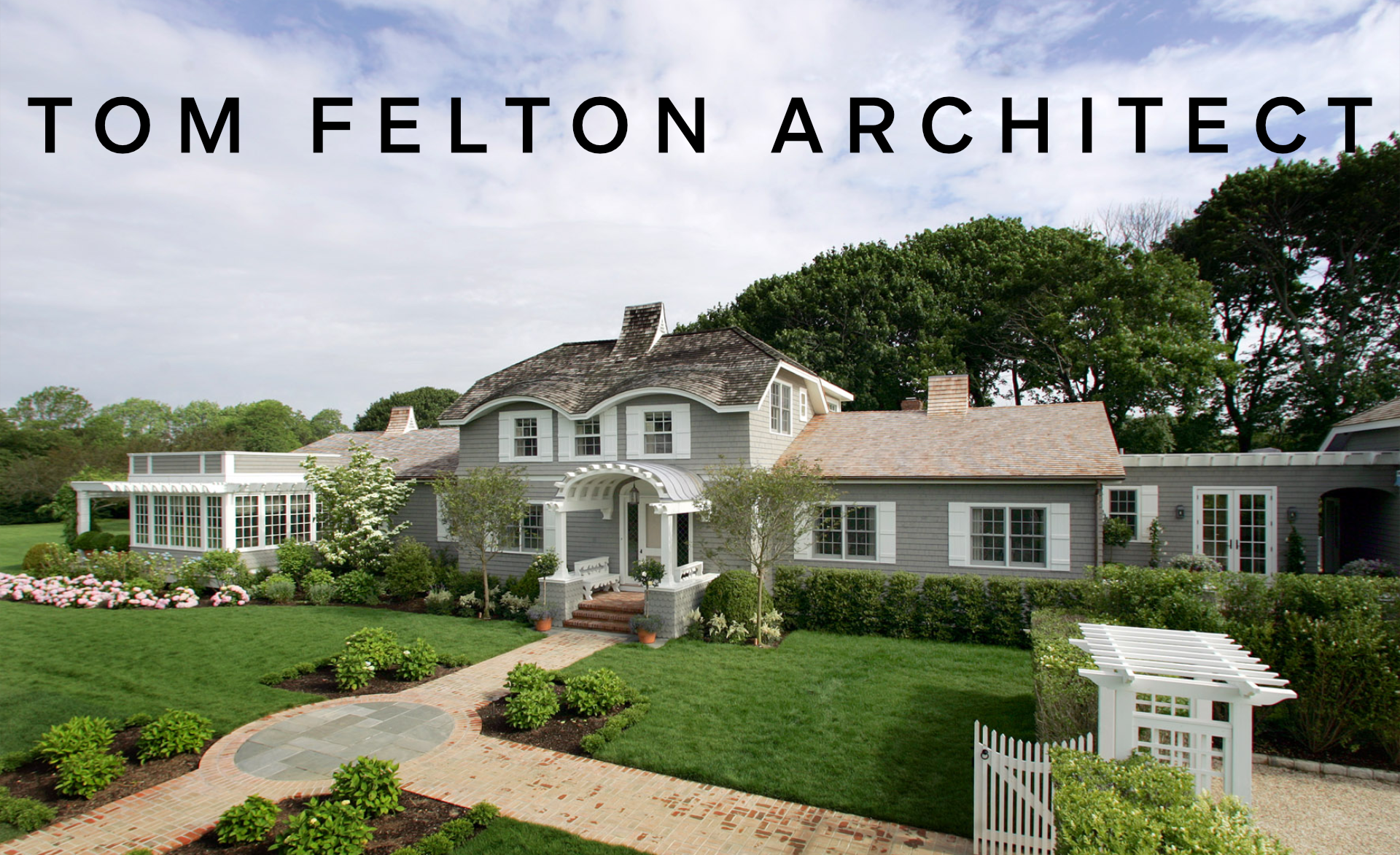 A large, gray, two-story house with a curved roof, surrounded by green lawn, trees, and flower beds, with a brick walkway leading to the front porch.