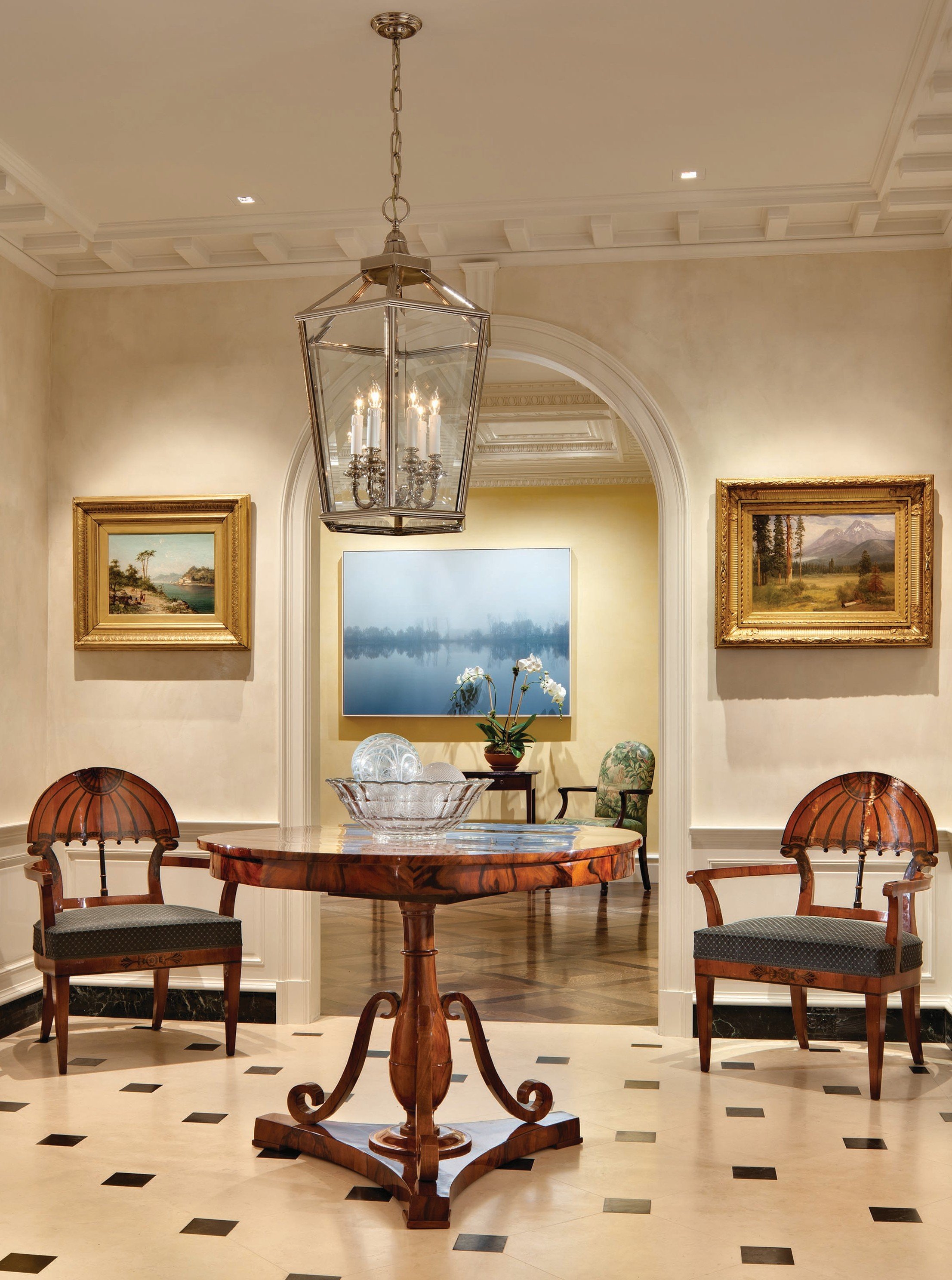 Elegant interior room with a wooden table, two wooden chairs, a chandelier, artwork on the walls, a potted plant, and an arched doorway leading to another room with additional furnishings and artwork.