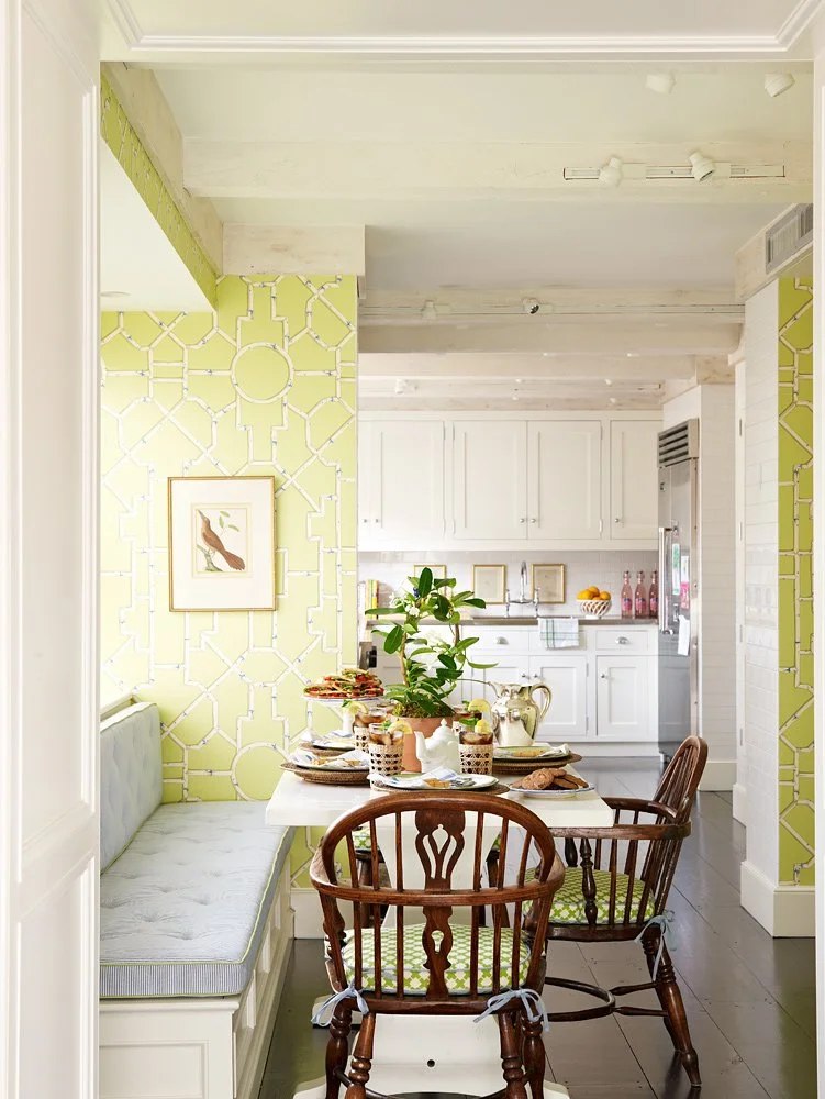 A cozy breakfast nook with a white table, wooden chairs, and a built-in cushioned bench against a decorative yellow patterned wall. The table is set with plates, teapots, cups, and a potted green plant. The background shows a white kitchen with cabin