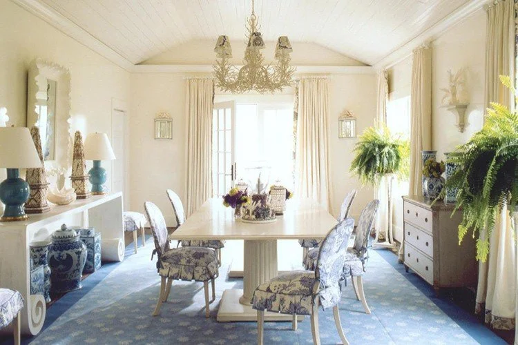 A spacious dining room with a long white table surrounded by upholstered chairs with floral patterns, light-colored curtains, a chandelier, decorative lamps, plants, and a sideboard with blue and white pottery.