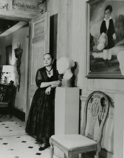 A woman in a long dark dress standing beside a pedestal with a bust sculpture, in an ornate room with paintings and antique furniture.