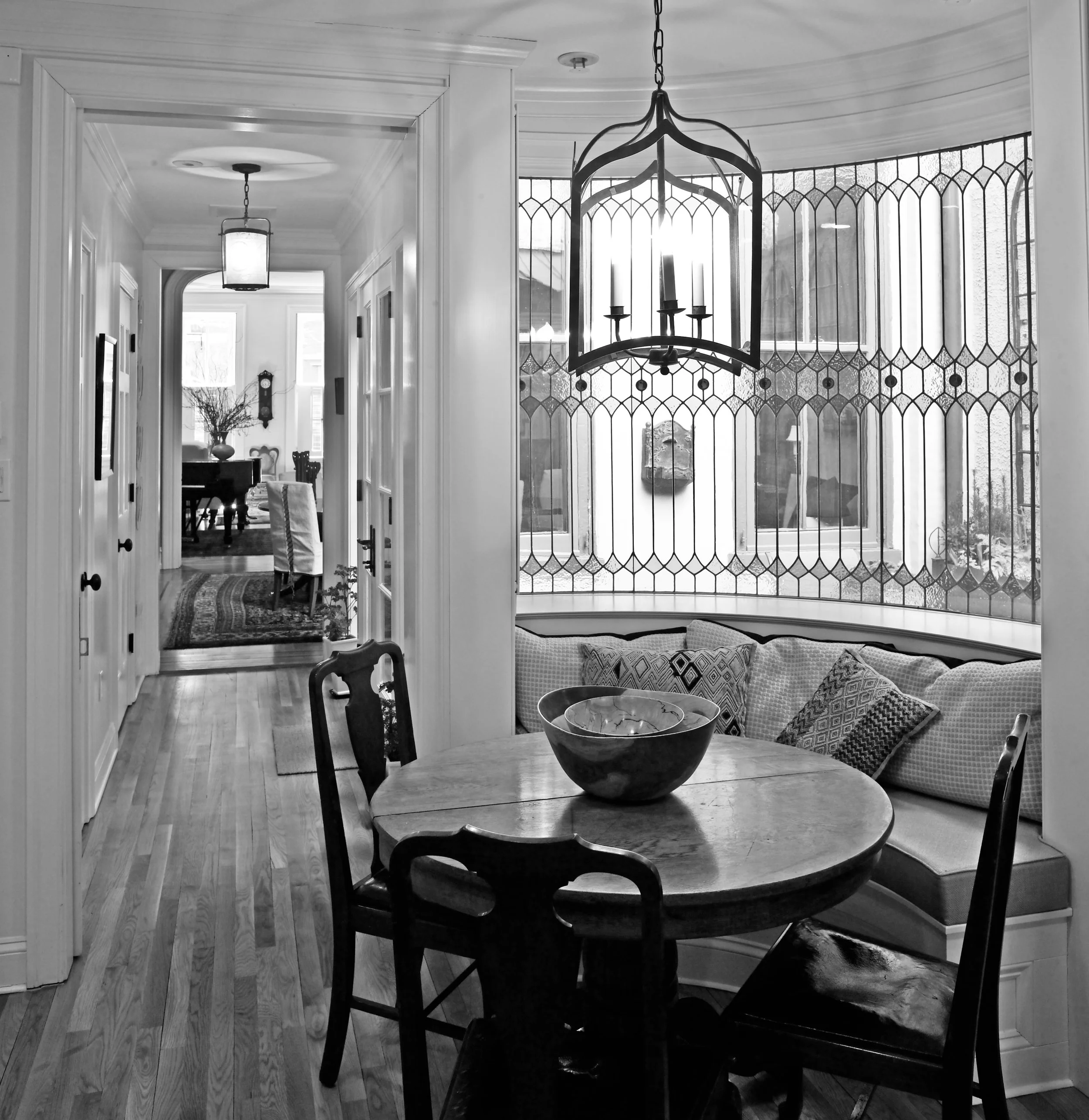 A black and white photo of a dining nook with a round table and chairs, overlooking a curved stained glass window with cushions along the window seat, hanging chandelier, and a view into a hallway leading to a living room.