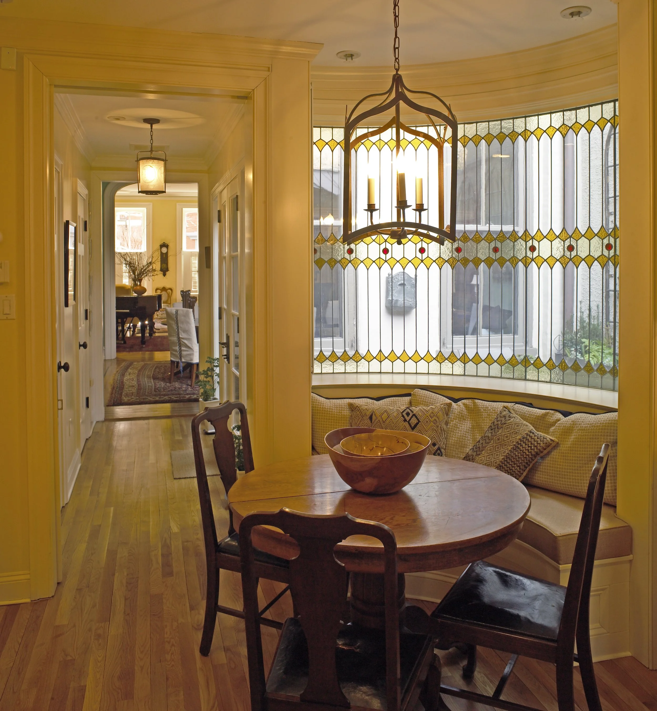 A cozy dining nook with a round wooden table, four chairs, and a built-in cushioned bench with patterned pillows. There is a large bowl on the table and a chandelier hanging above. A stained glass window with jewel-tone accents lets in natural light,