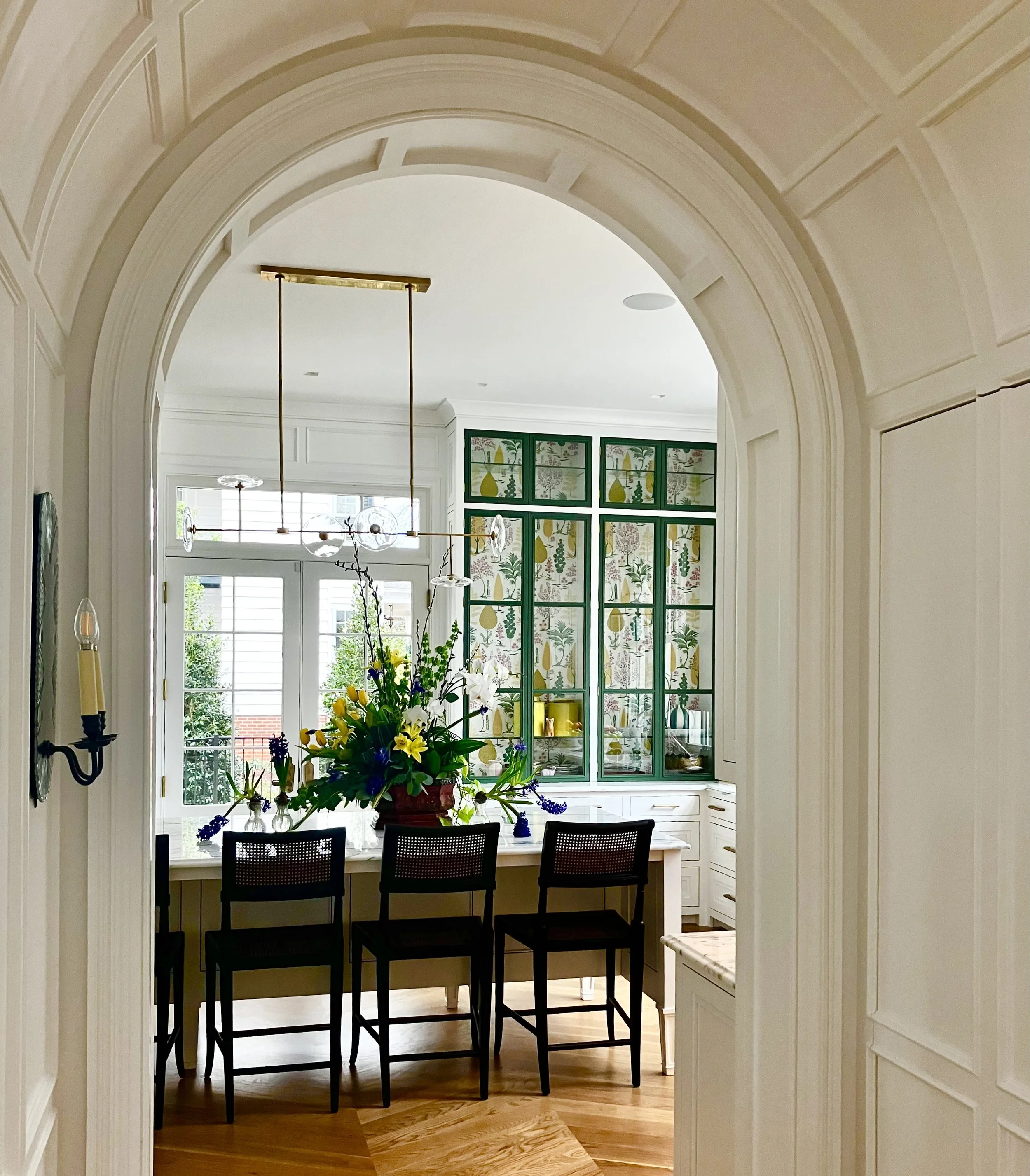 View through an archway into a bright dining area with a large floral centerpiece on a white table, modern black chairs, and green framed glass cabinet doors with decorative wallpaper inside.