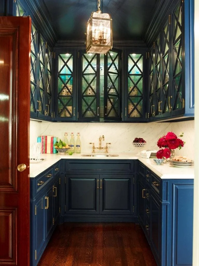 A small kitchen with dark blue cabinets, a white marble countertop, and brass hardware. Above, glass-front cabinets with X-shaped dark blue framing. A hanging lantern-style light fixture illuminates the space. The counter has colorful books, bottles,