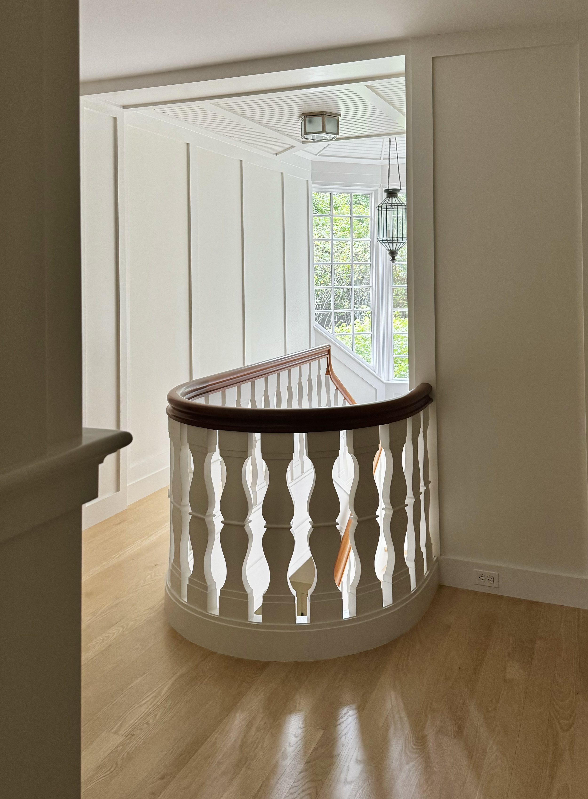 Interior view of a house staircase with white balustrades, a wooden handrail, and large windows letting in natural light.