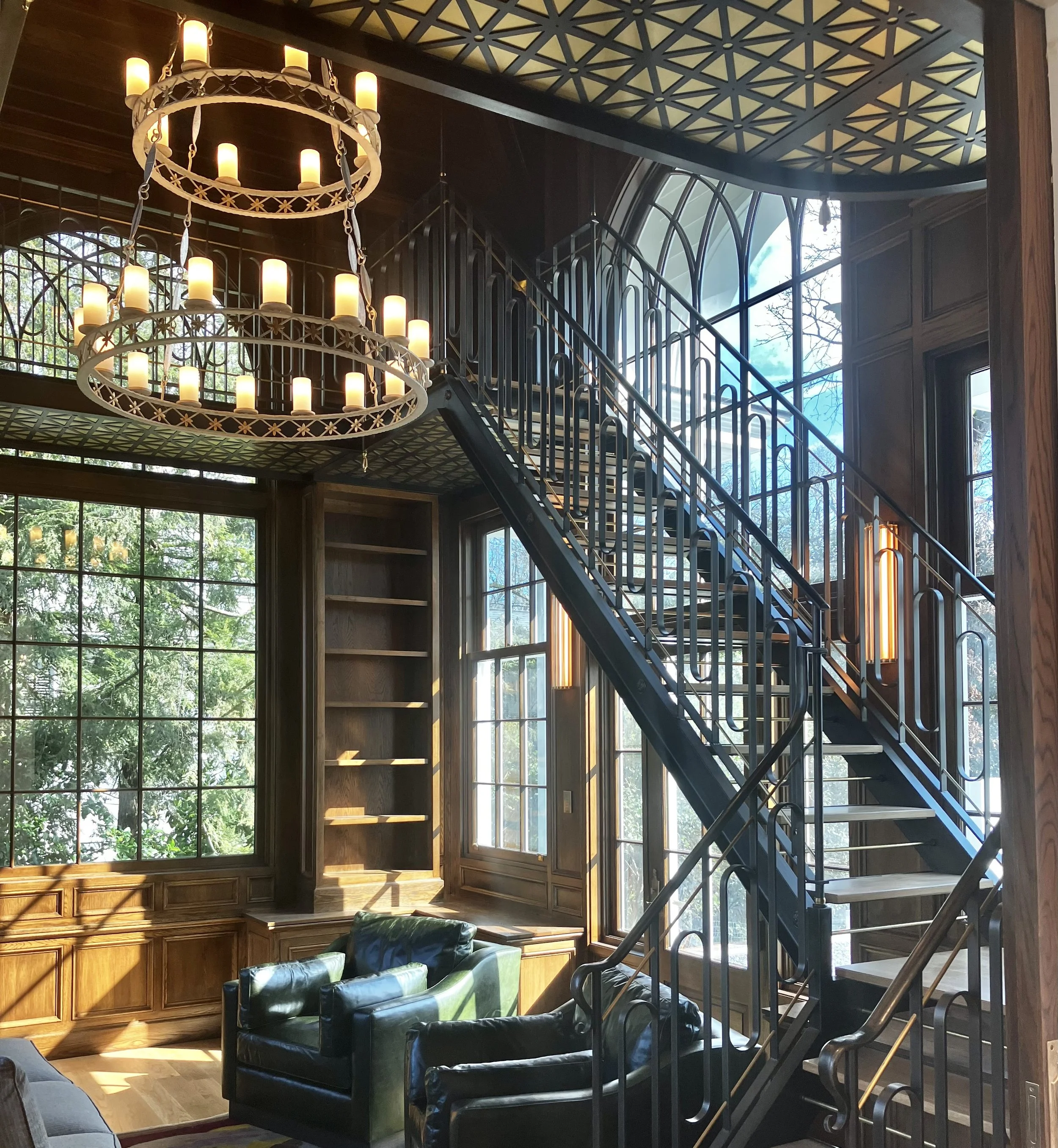 Interior of a two floor residential library with large arched windows, a metal staircase, a large chandelier with candle-shaped lights, wooden bookshelves, and leather chairs, illuminated by natural sunlight.