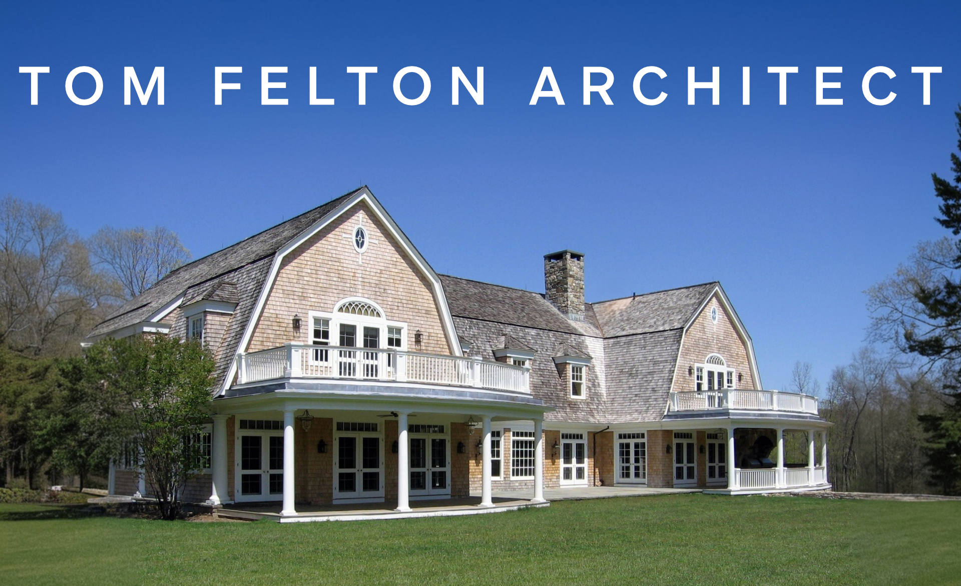 A large, two-story house with a shingled roof, multiple balconies, and white columns on a lush green lawn under a bright blue sky.
