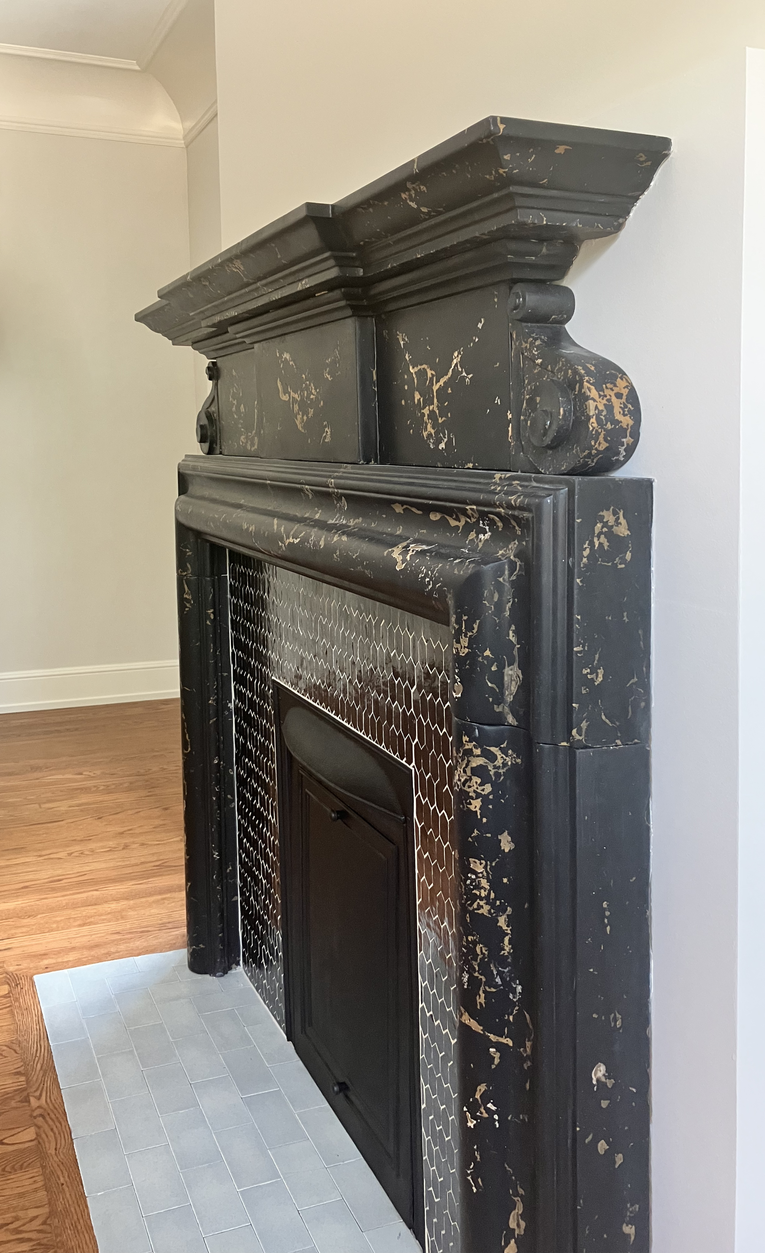Close-up of an antique black fireplace with distressed finish, honeycomb patterned tile around the opening, situated in a room with hardwood flooring and white walls.