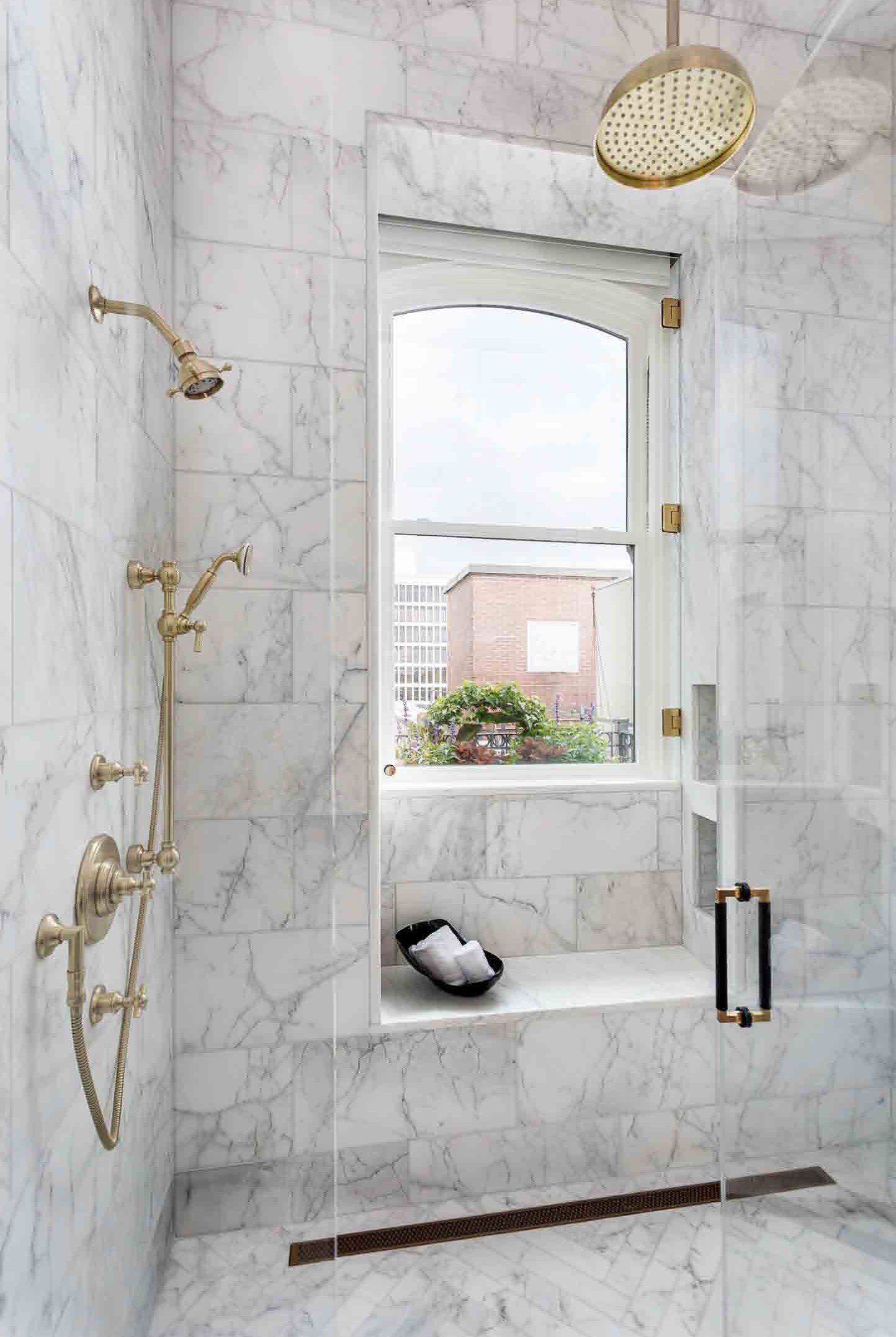 A marble shower with a window, a brass showerhead, and a built-in shelf with a black bowl holding towel. Outside view through the window shows a building and foliage.