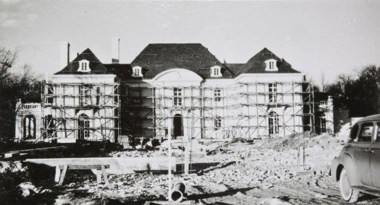 Black and white photo of a large, historic mansion under construction, surrounded by scaffolding, with a vintage car parked nearby.