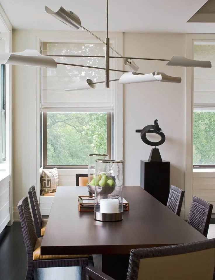 Modern dining room with a dark wood table, six wicker chairs, a centerpiece with green apples in glass jars, a white candle on a silver base, and abstract black sculpture on a pedestal. Large windows with white blinds letting in natural light.