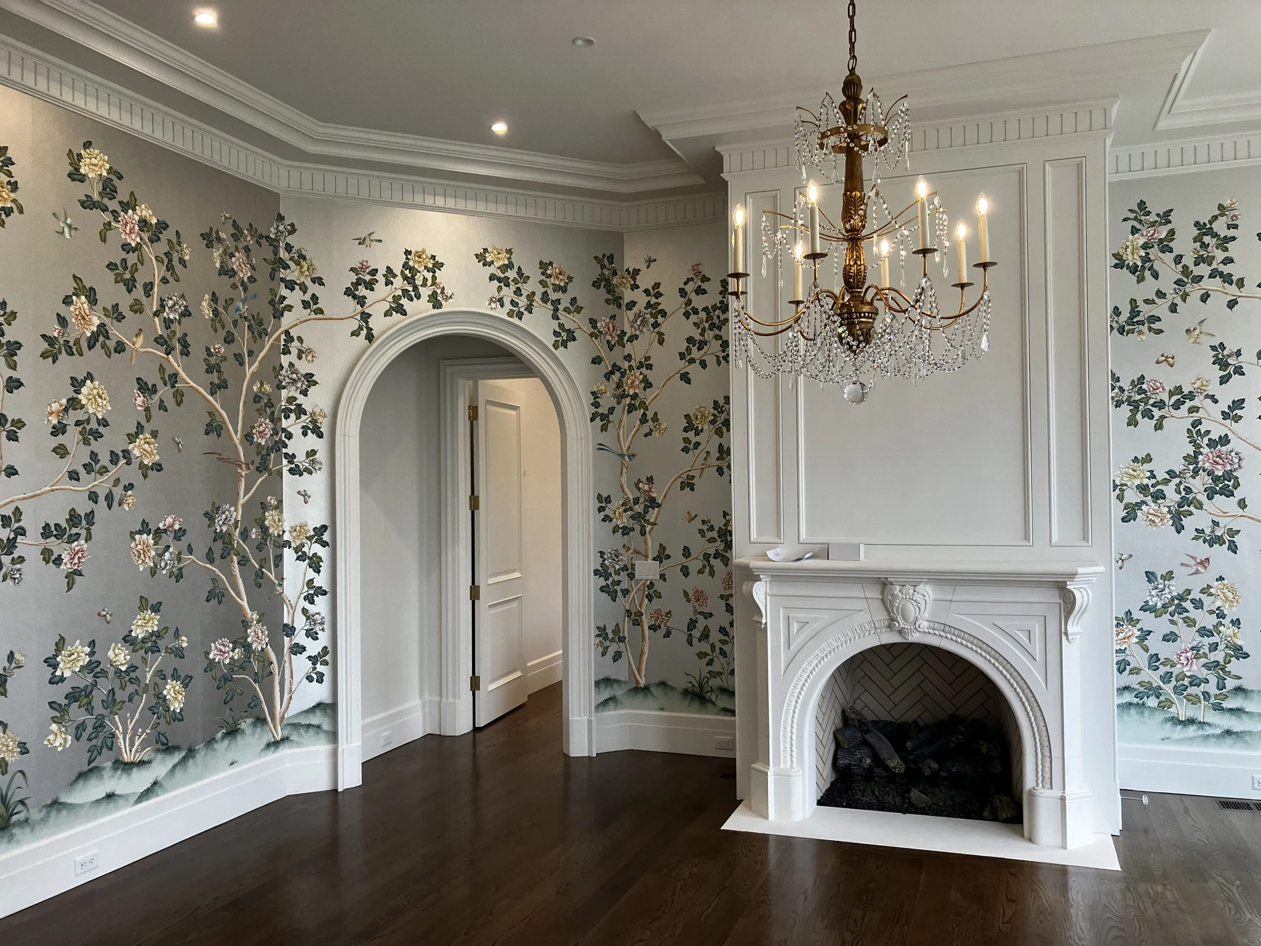 A room with floral wallpaper, a white fireplace, and a gold chandelier with candle-style lights and hanging crystals.