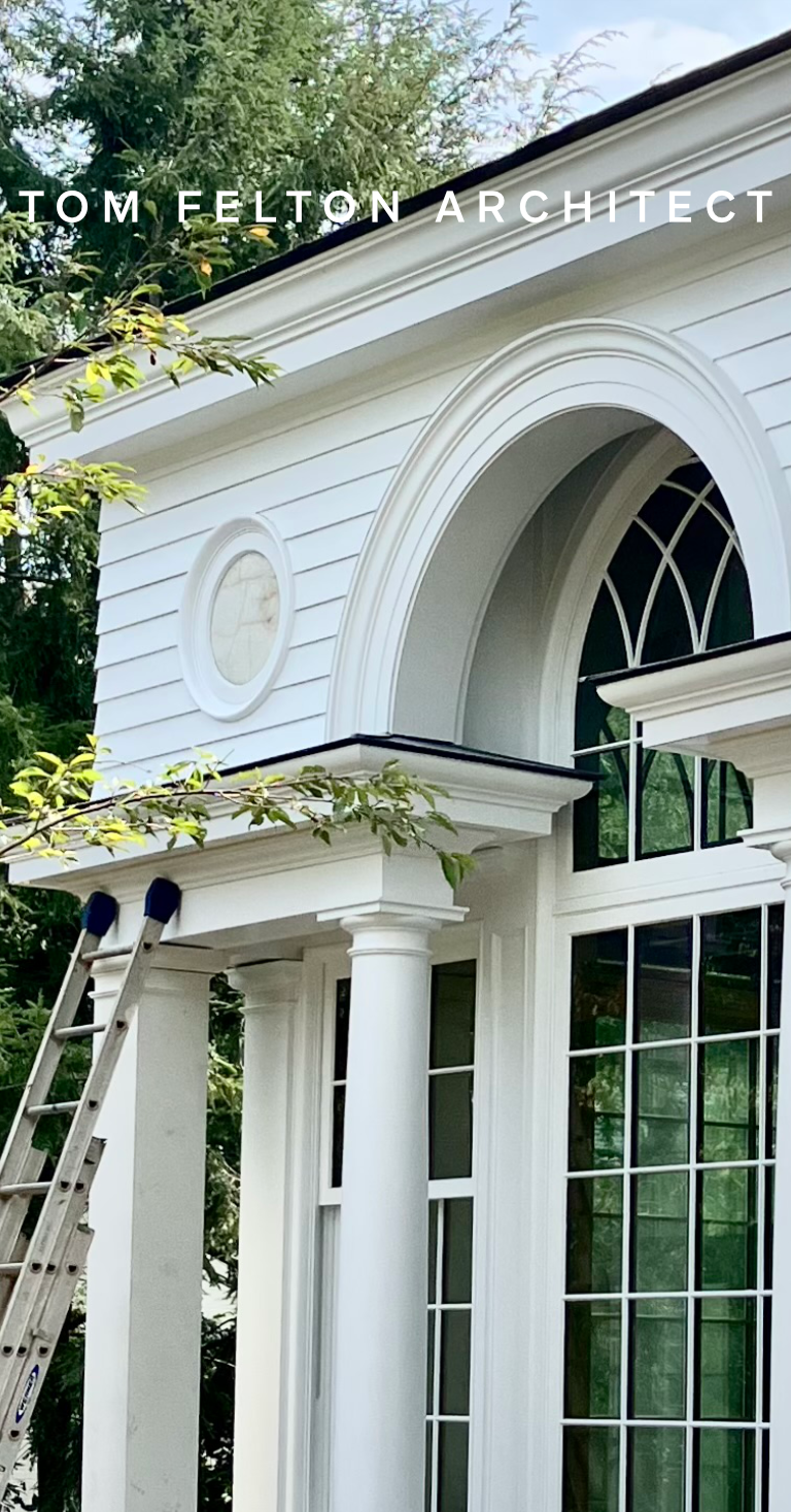 Close-up of a white house's exterior with large arched window, decorative trim, and classical column, surrounded by trees.