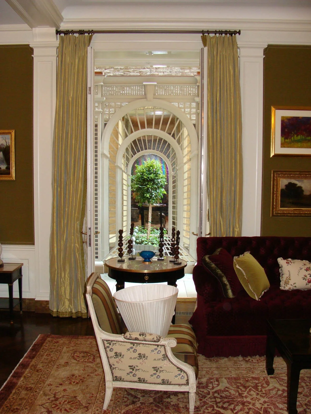 Living room with an open door leading to a small patio area with a potted tree, decorated with arches and latticework, and furnished with various chairs, a sofa, and framed artwork on the walls.