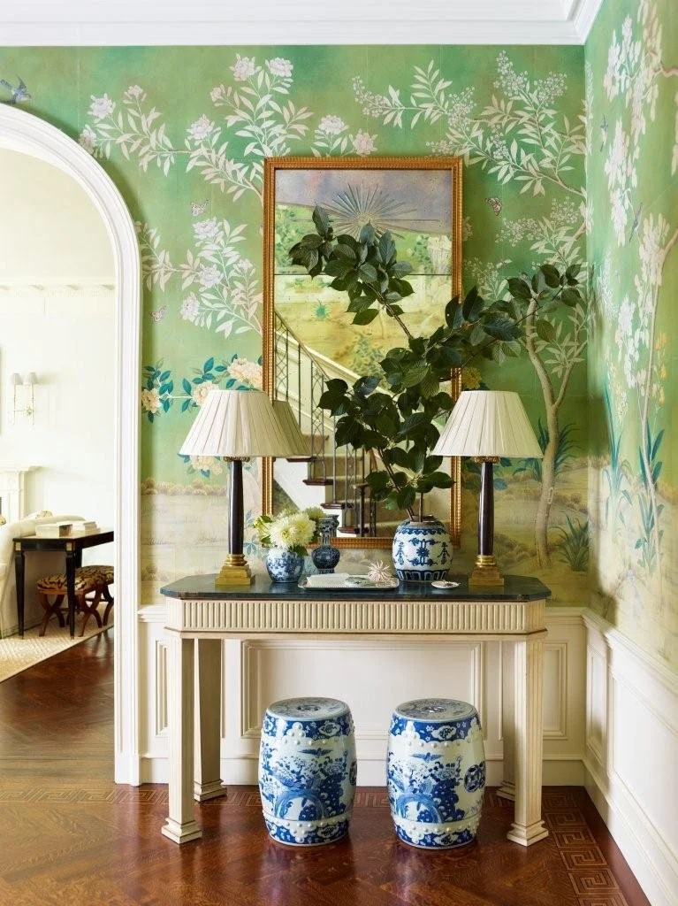 A hallway with a green floral wallpaper, a white console table topped with two lamps, a large plant, and decorative blue and white porcelain stools underneath. A mirror above the table reflects a staircase.