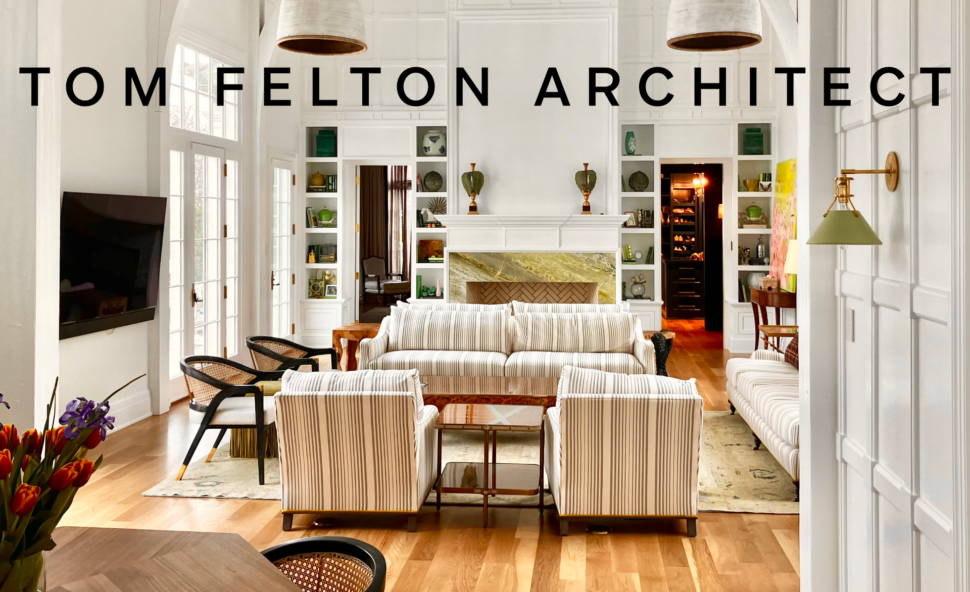 Elegant living room with white paneled walls, a marble fireplace, and built-in shelves filled with decor. Light-colored striped sofas and armchairs are arranged around a wooden coffee table. French doors let in natural light, and a flat-screen TV is 