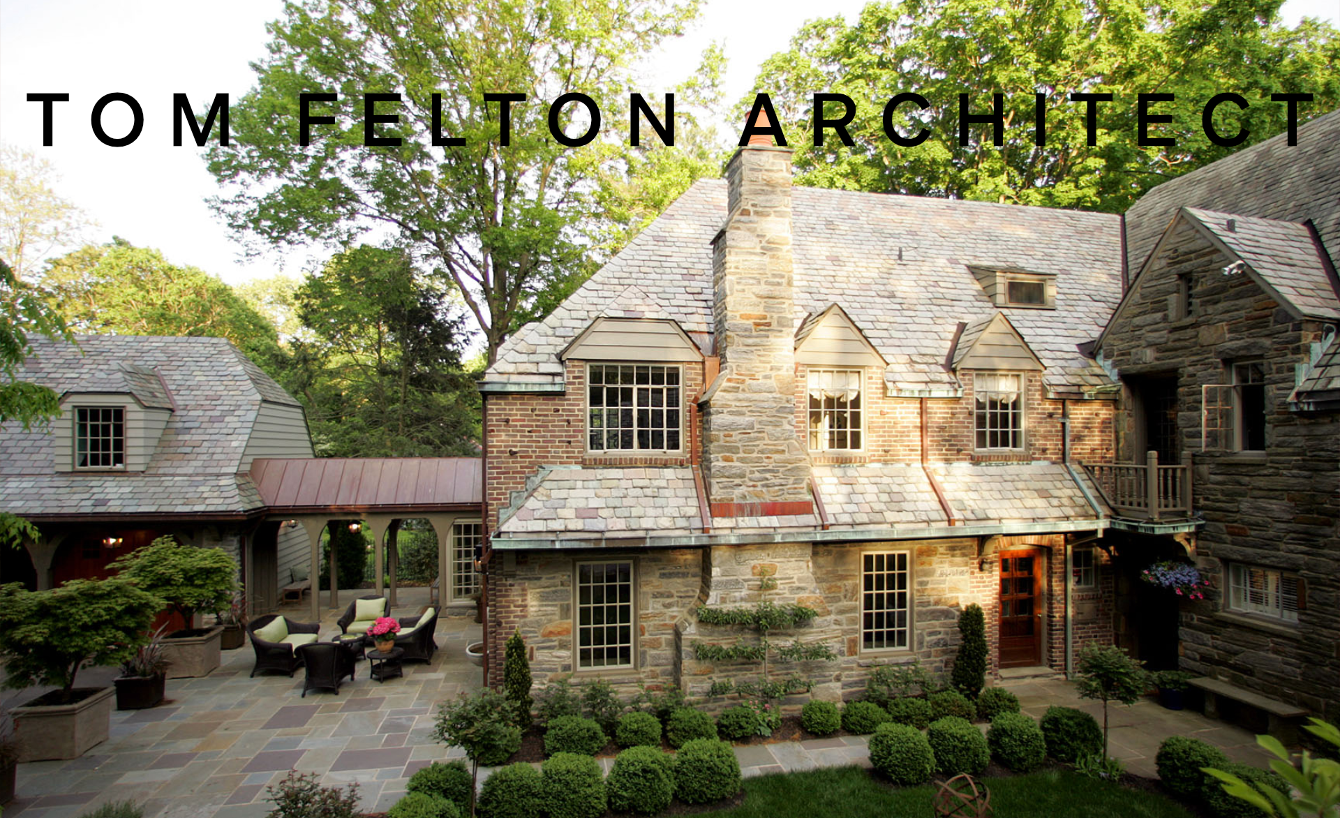 Exterior view of a stone and brick house with a slate roof, surrounded by green trees, and a patio with outdoor seating and potted plants.