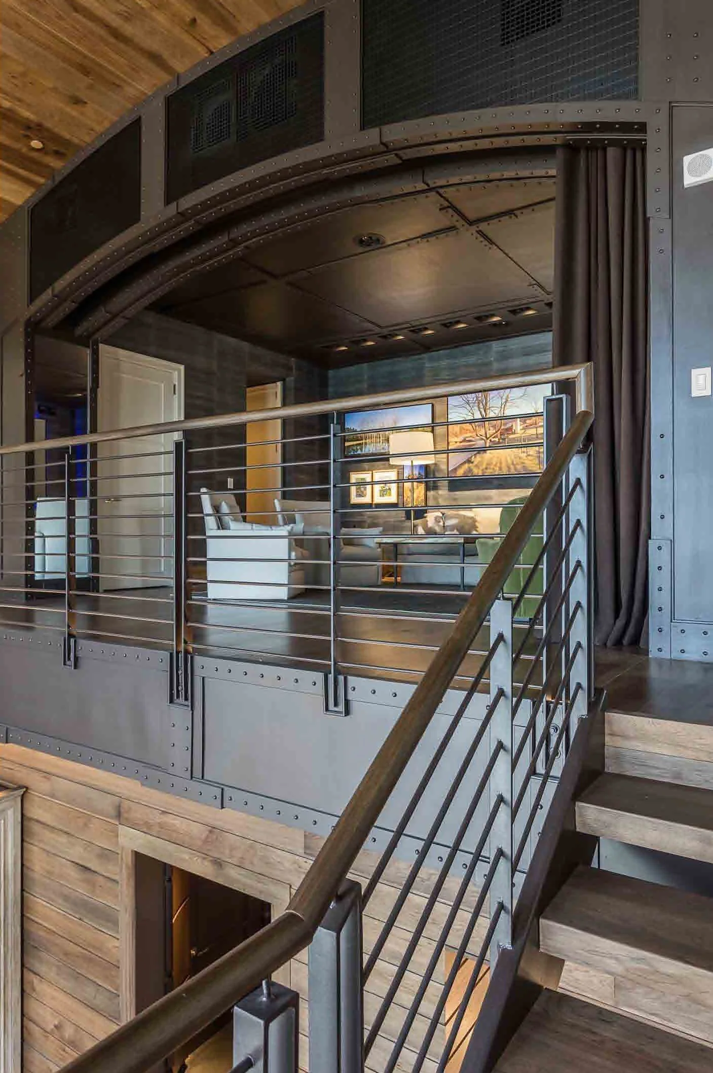 Interior view of a modern home with wooden staircase, metal railing, and a loft area overlooking a living room with white furniture and wall art.