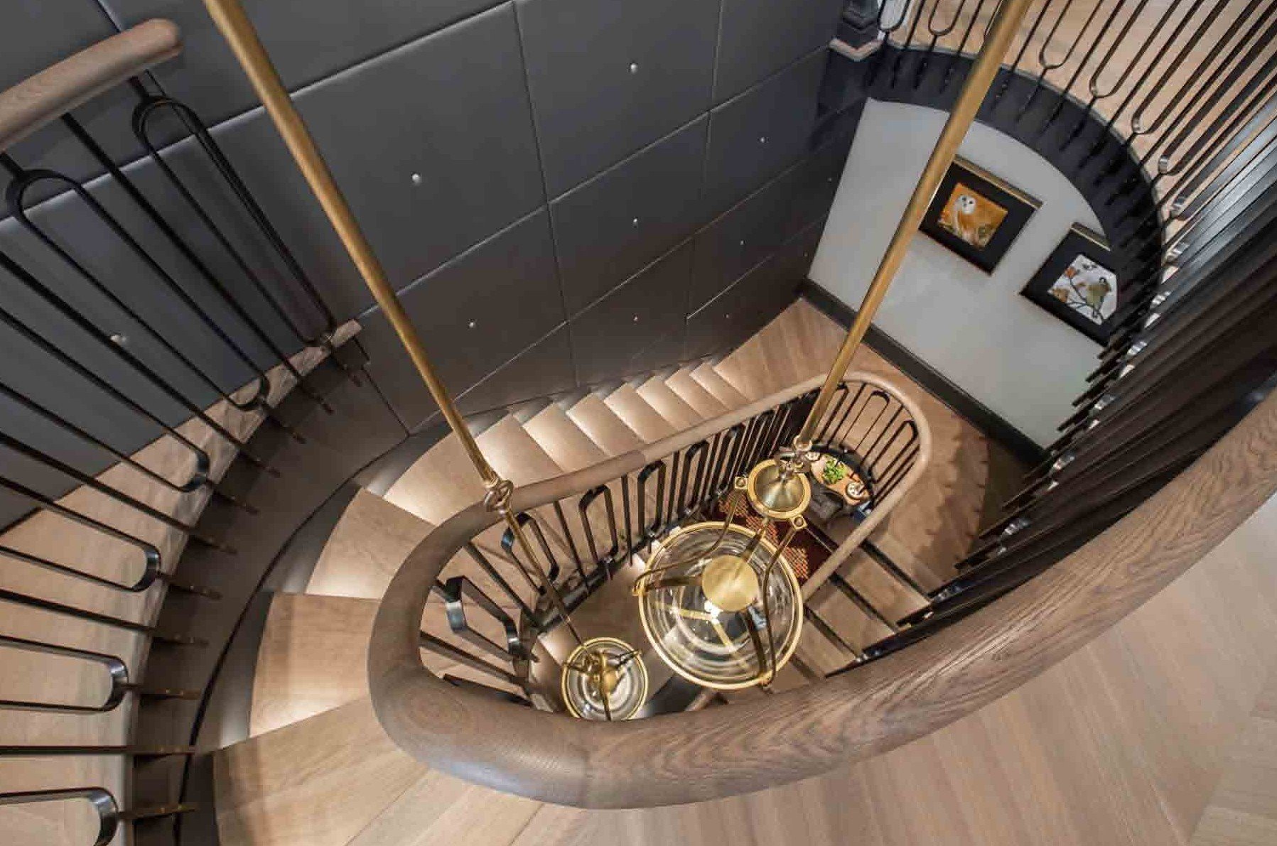 A top-down view of a spiral staircase with wooden steps, black metal railing, and a brass chandelier hanging in the center. The staircase surrounds a wall decorated with framed artwork.