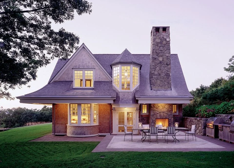 A large house with a steep roof, stone chimney, and a spacious backyard patio with outdoor seating and a built-in grill.