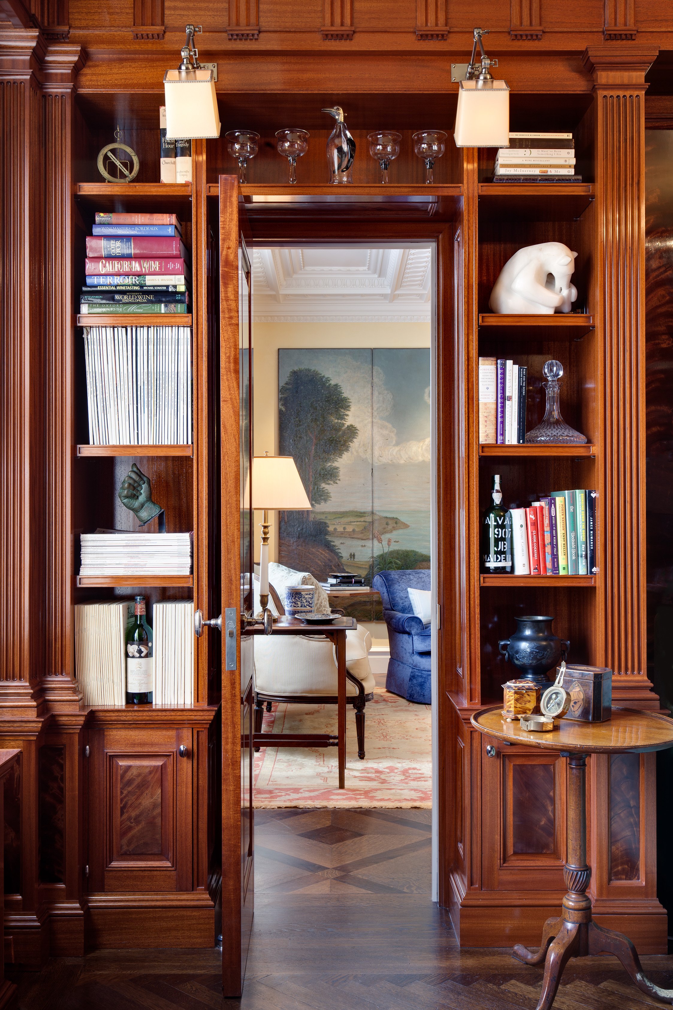 View through a wooden doorway revealing a cozy living room with a painting of a landscape, a blue armchair, a beige armchair with a lamp, and various decorative objects and books on the shelves.