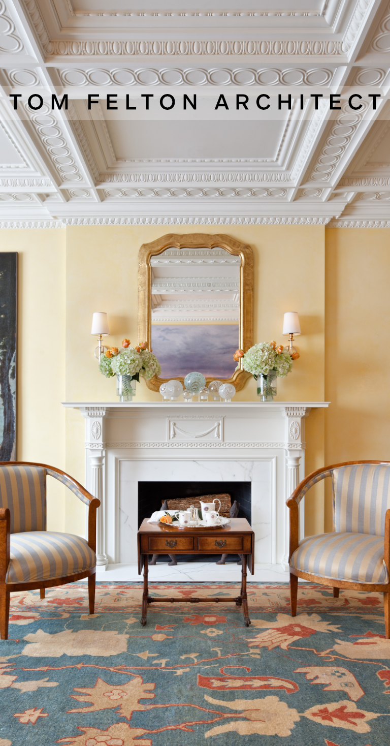 A cozy living room with a white fireplace mantel, a gold-framed mirror, two wall-mounted lamps, decorative vases and flowers, two striped armchairs, a small wooden table set with a tea set, and an ornate rug on the floor.