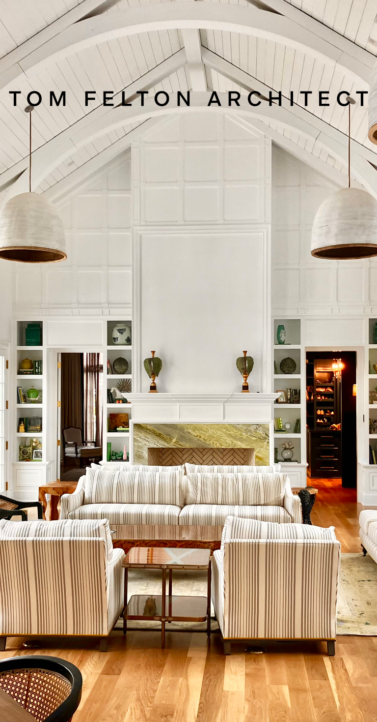 Living room with white vaulted ceiling, built-in shelves, striped sofa and armchairs, fireplace with gold and marble accents, and two large hanging pendant lights.