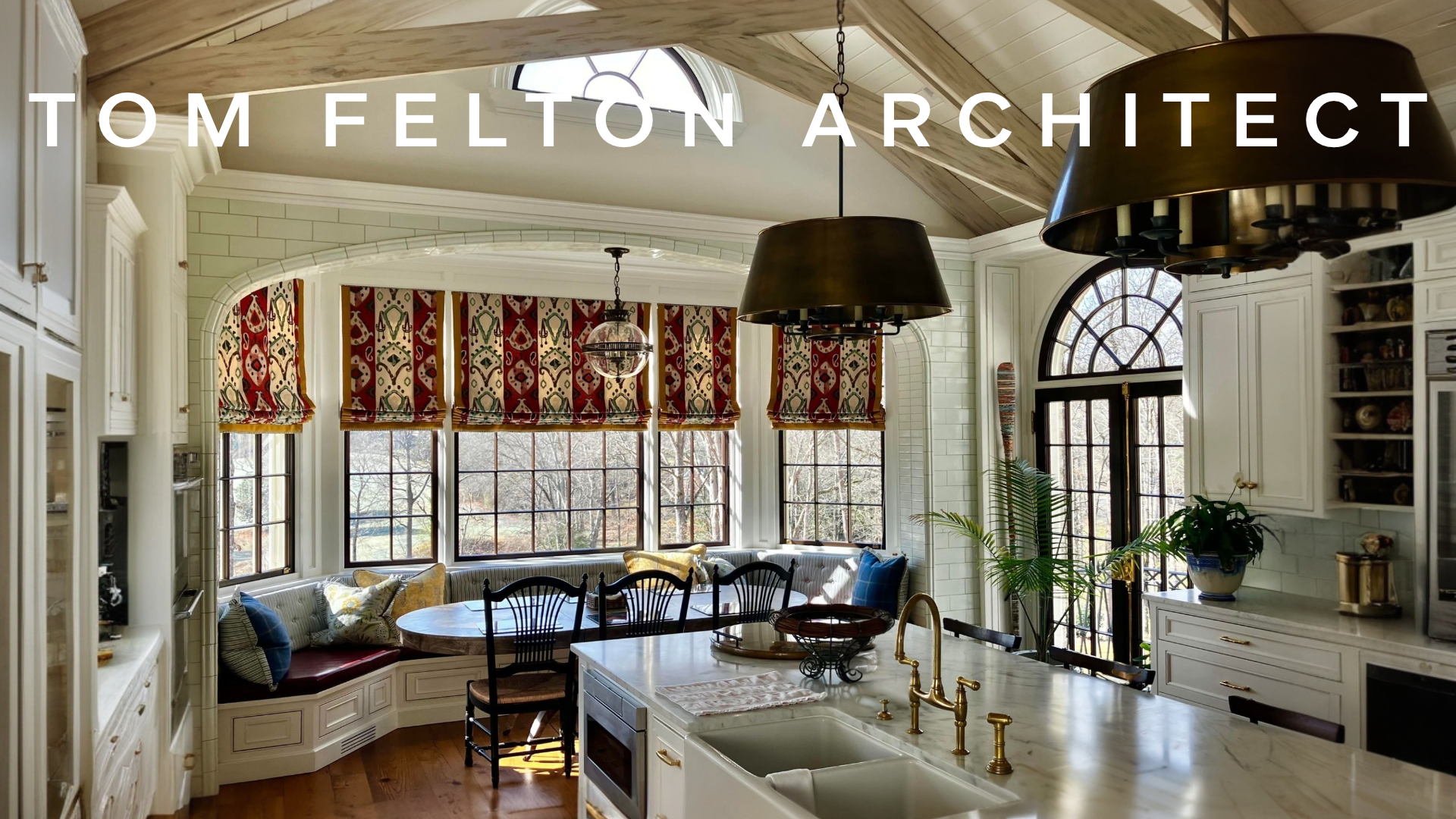 Bright kitchen with white cabinets, a marble island with a sink and gold fixtures, a built-in window seat with cushions, large windows with patterned red, beige, and green Roman shades, and a skylight.