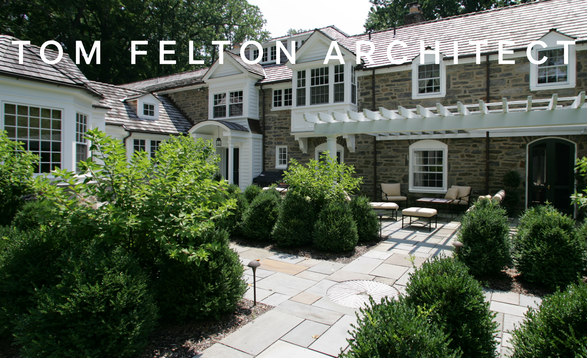 Front yard of a large stone house with white trim, surrounded by landscaped bushes and patio furniture.