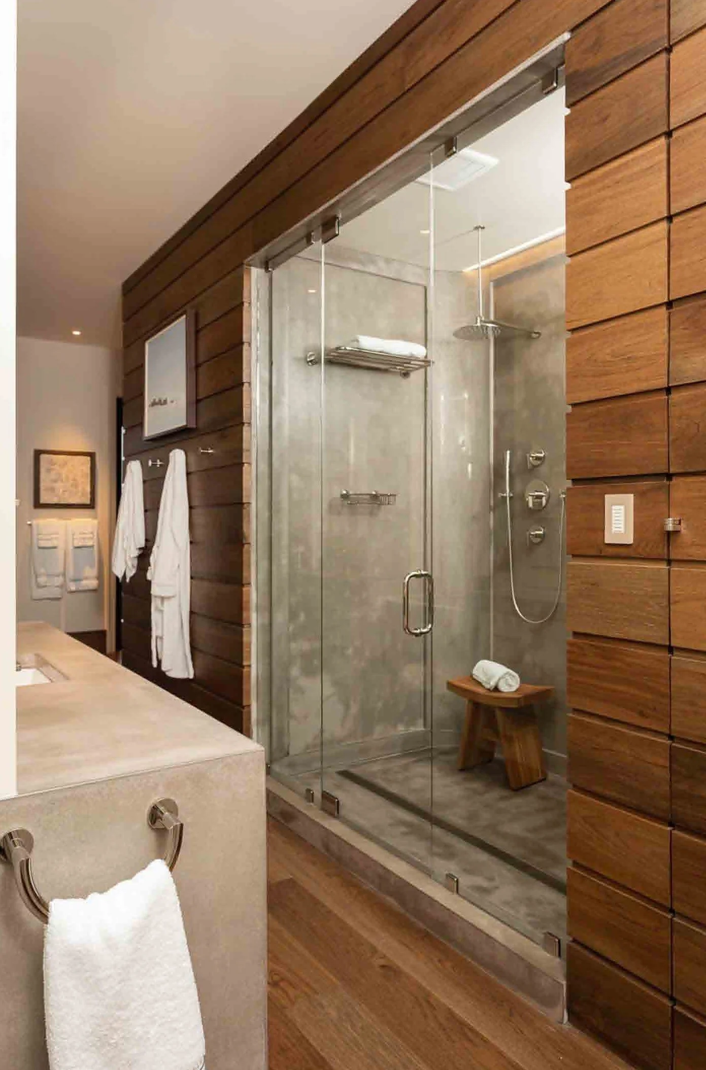 Modern bathroom with a glass-enclosed shower, wooden wall, towel rack, white towels, and a small bench with a rolled towel.