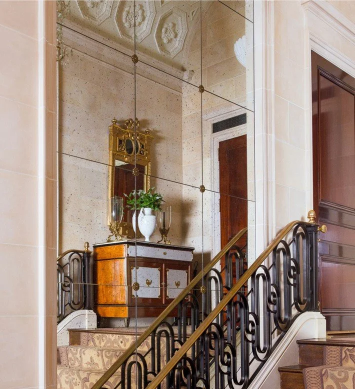 A mirrored wall in a luxurious hallway reflects a wooden sideboard with a white vase holding green foliage, a gold-framed mirror, and candles. The staircase has ornate black iron railings with gold accents, and the ceiling features decorative molding