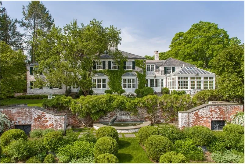 Large house with multiple floors, gray exterior, numerous windows, dining room with a glass ceiling, surrounded by green trees and bushes, with a landscaped backyard featuring a fountain and brick walls.