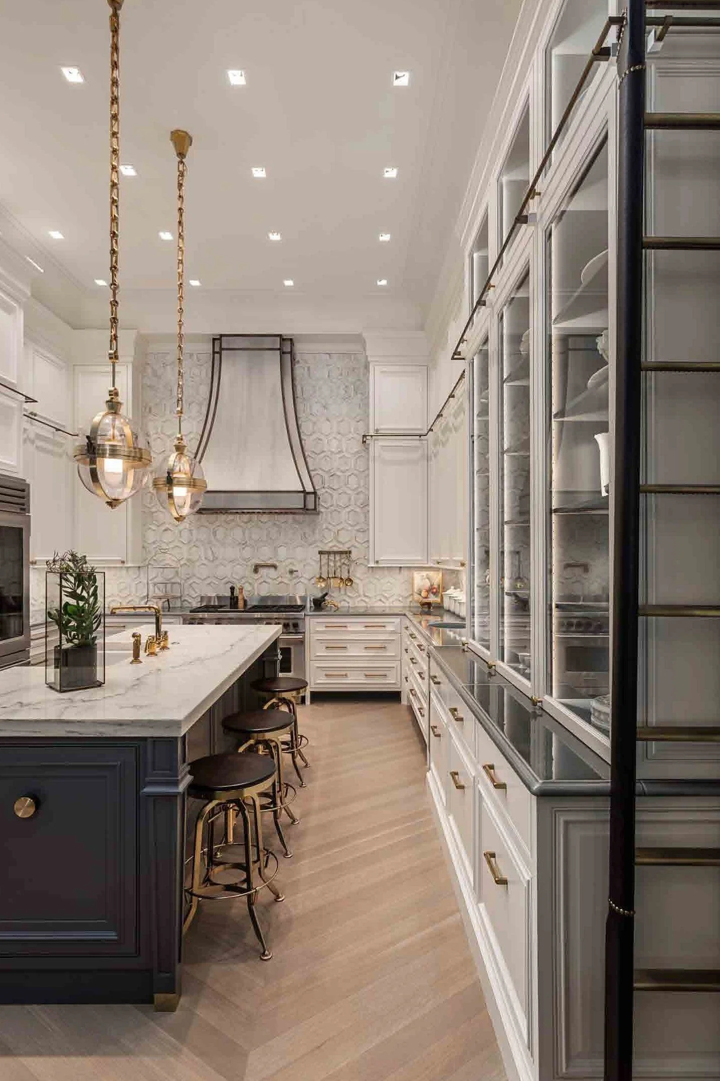 Modern kitchen with white cabinetry, a blue island with a marble countertop, and brass accents. Two pendant lights hang over the island, and there is a glass-front pantry on the right side.