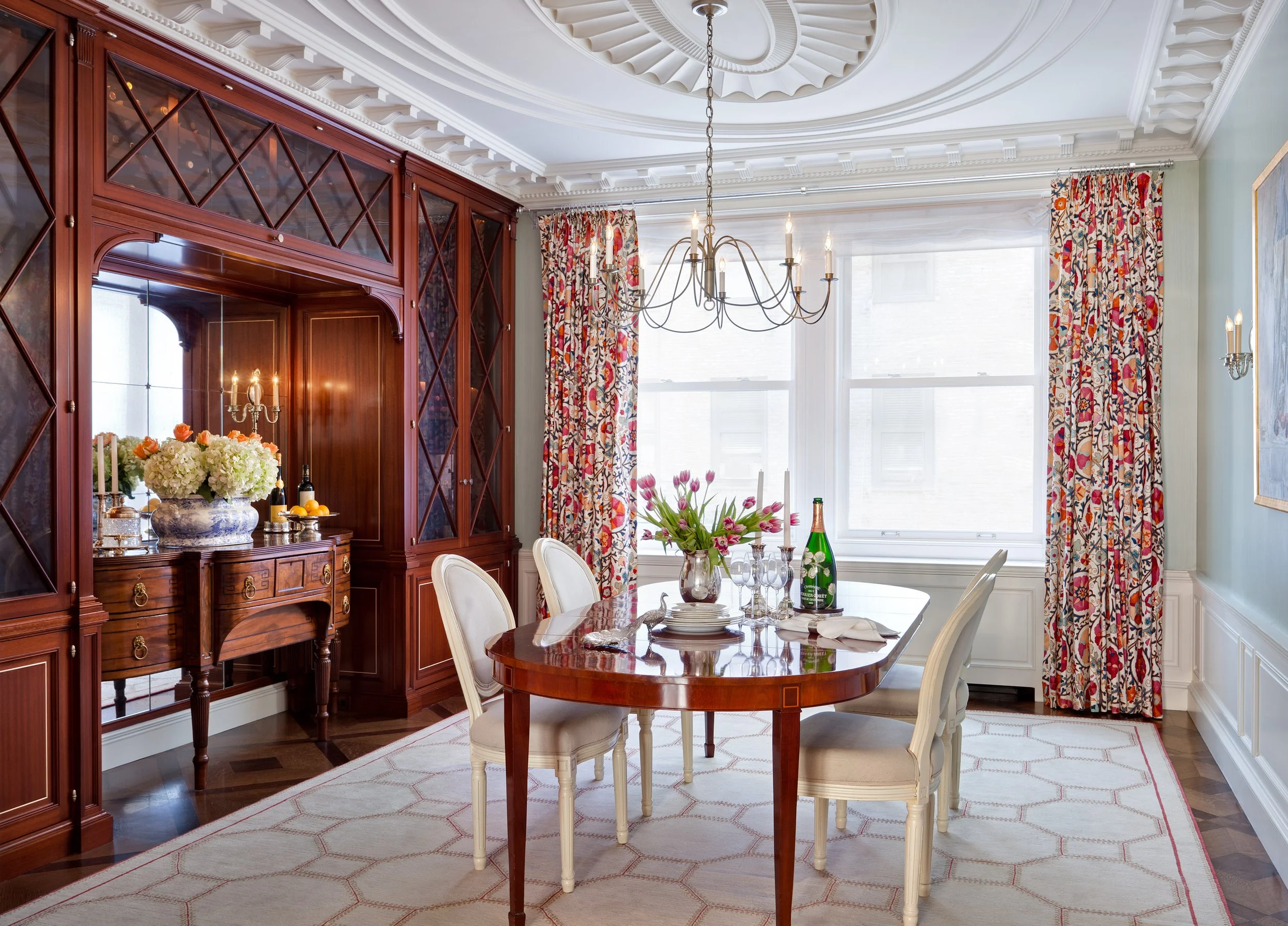 Elegant dining room with a polished wooden table set with plates, glasses, and a floral centerpiece. The room features large windows with patterned curtains, a chandelier hanging from the ceiling, and a wooden china cabinet on the left side, all with