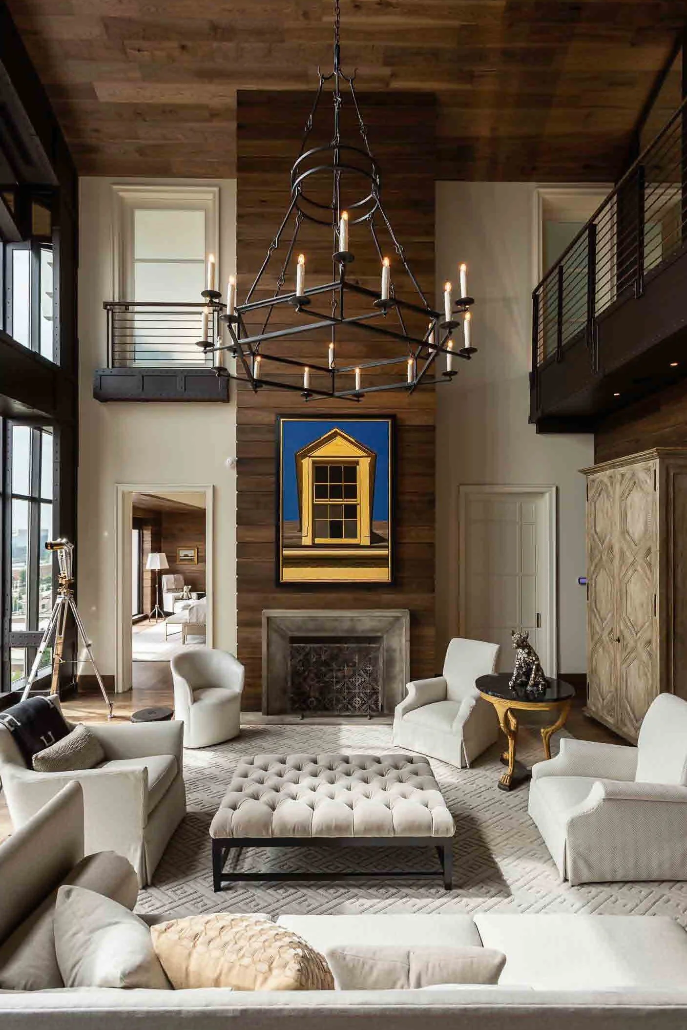 A spacious living room with a high wooden ceiling, large windows, and a black iron chandelier. There are white armchairs and a tufted lounge ottoman arranged around a fireplace with a colorful framed painting above it. The room features wooden accent