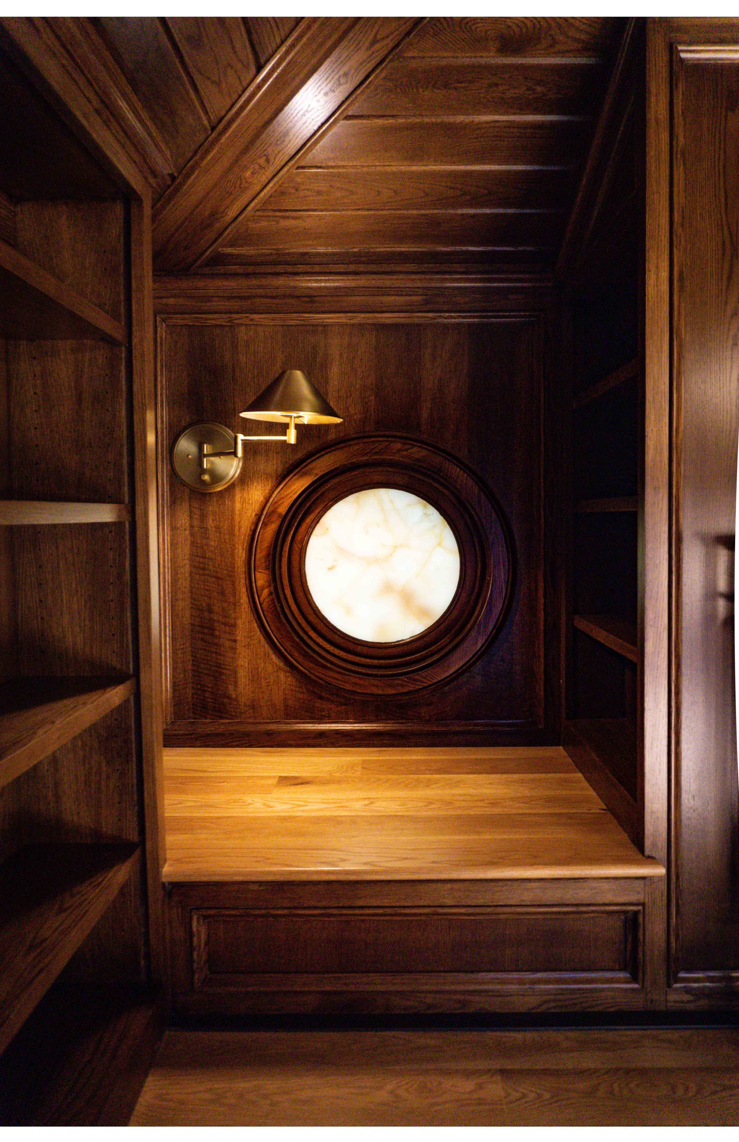 A wooden nook with built-in shelves on the left and right, a small brass wall lamp, and a round, backlit marble window on the back wall.