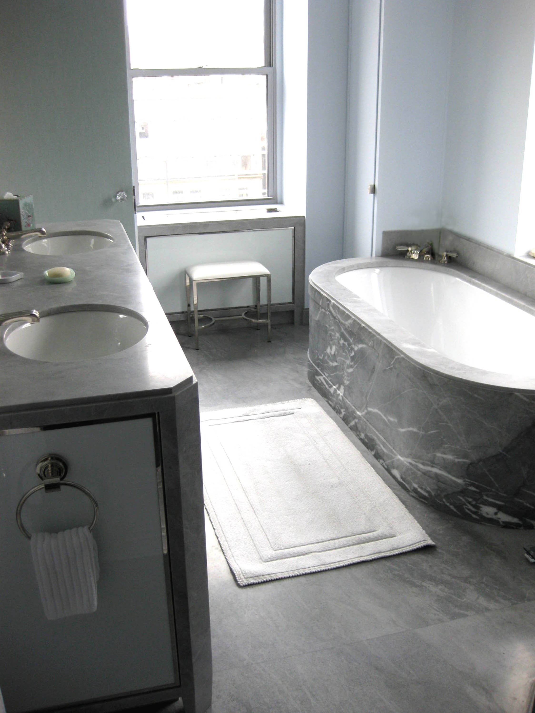 A modern bathroom with a large bathtub, a double sink vanity, a window, and a white rug on gray tile flooring.