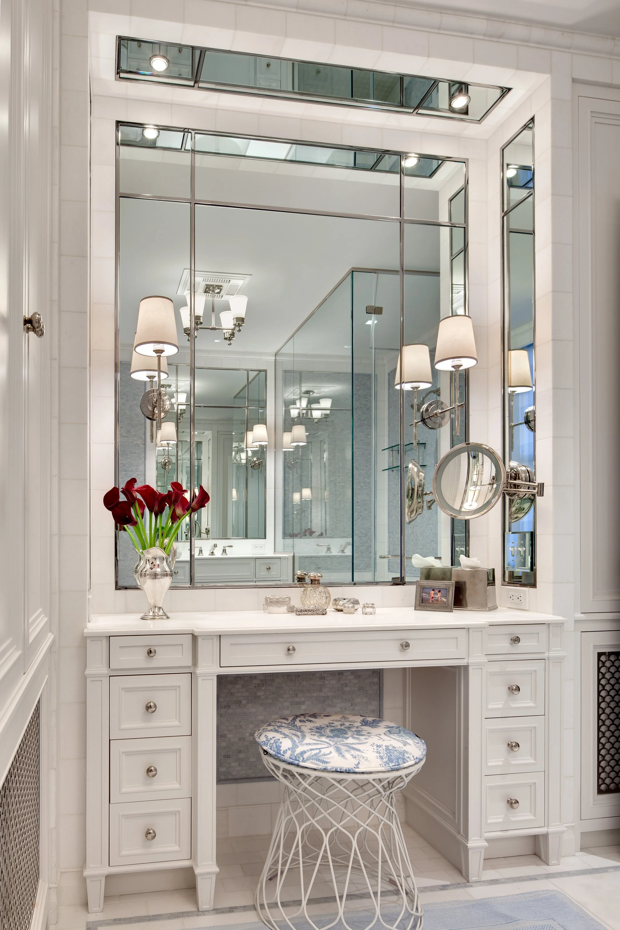 A white vanity with a large mirror, decorative accessories, and a vase of red calla lilies, in a bright bathroom with multiple wall-mounted lamps and a cushioned stool with a blue and white floral pattern.