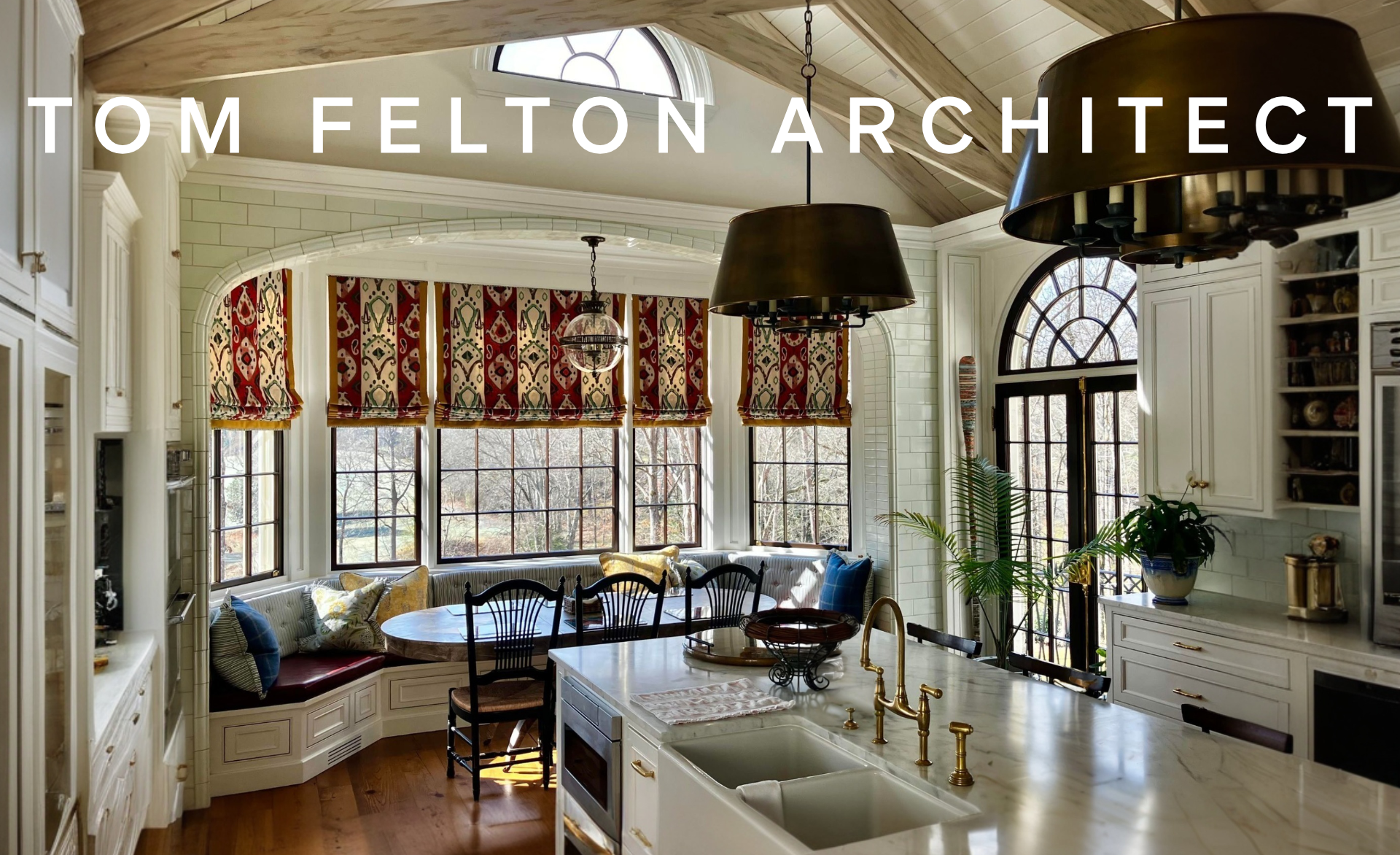Interior view of a bright kitchen with a curved window nook finished in tile, featuring a round table and cushioned seating, multiple windows with patterned Roman shades, white cabinetry, a large island with a double sink and gold fixtures.