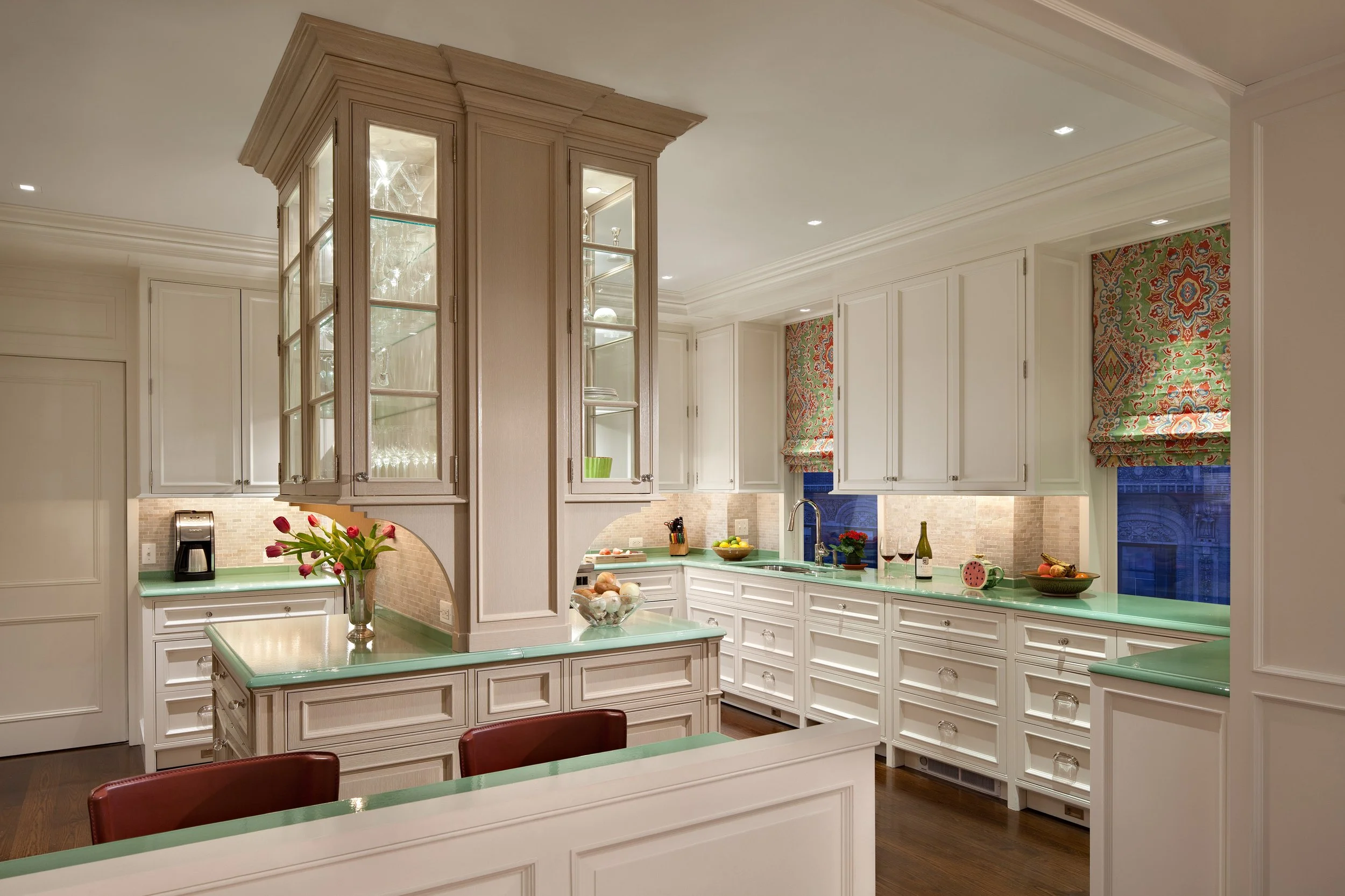 Bright white kitchen with green countertops, glass-front cabinets, and colorful window valances. Decorated with flowers, wine, and kitchen essentials.