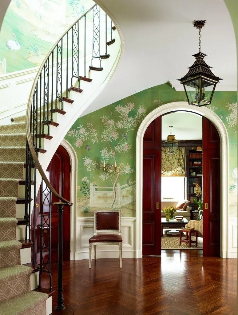 Entryway of a house featuring a curved staircase with a dark iron railing, green decorative wallpaper with floral and bird motifs, a wall-mounted sconce, a chair with black leather upholstery, and an arched doorway leading to a sitting room with wood