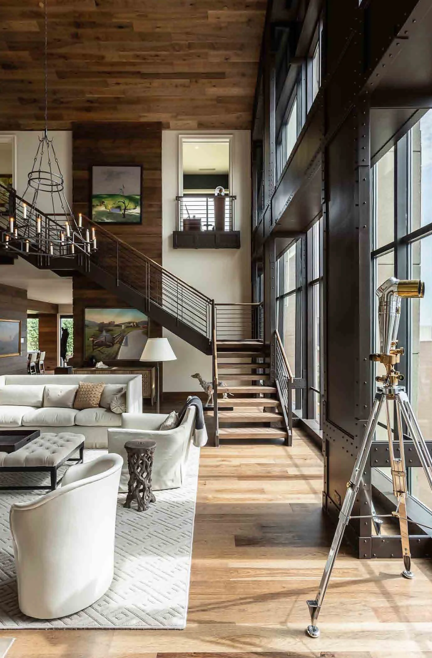 Interior of a modern house with wooden floors and walls, a white sectional sofa, a telescope on a tripod, and large floor-to-ceiling windows with metal framing.