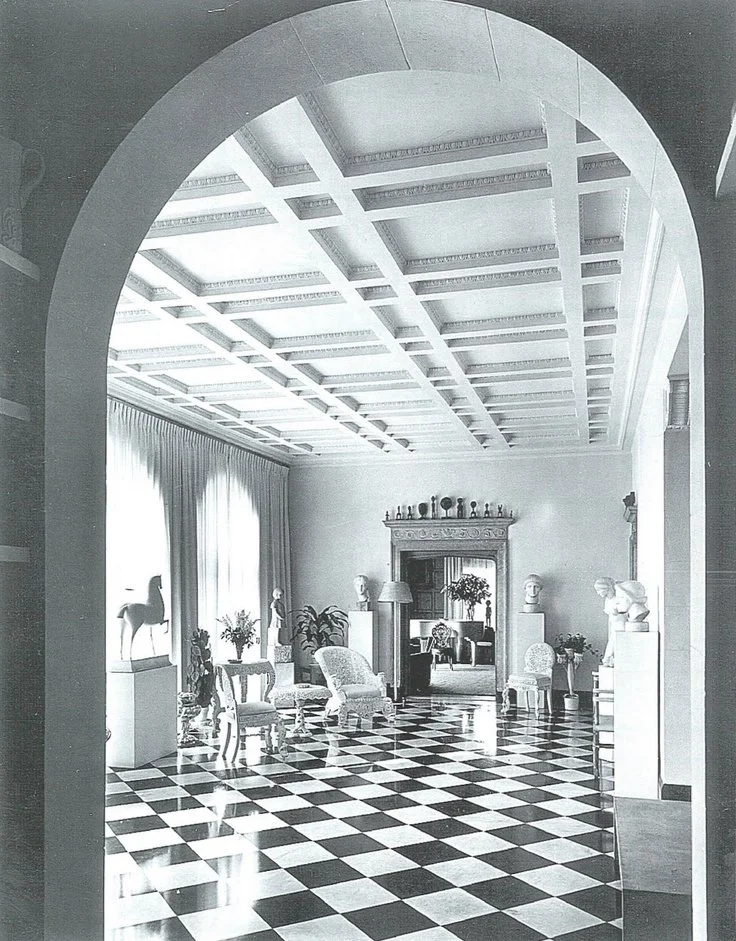 A grand, elegant interior room with checkered black and white floor, ornate ceiling, large windows with drapes, and classical sculptures and plants, viewed through an archway.