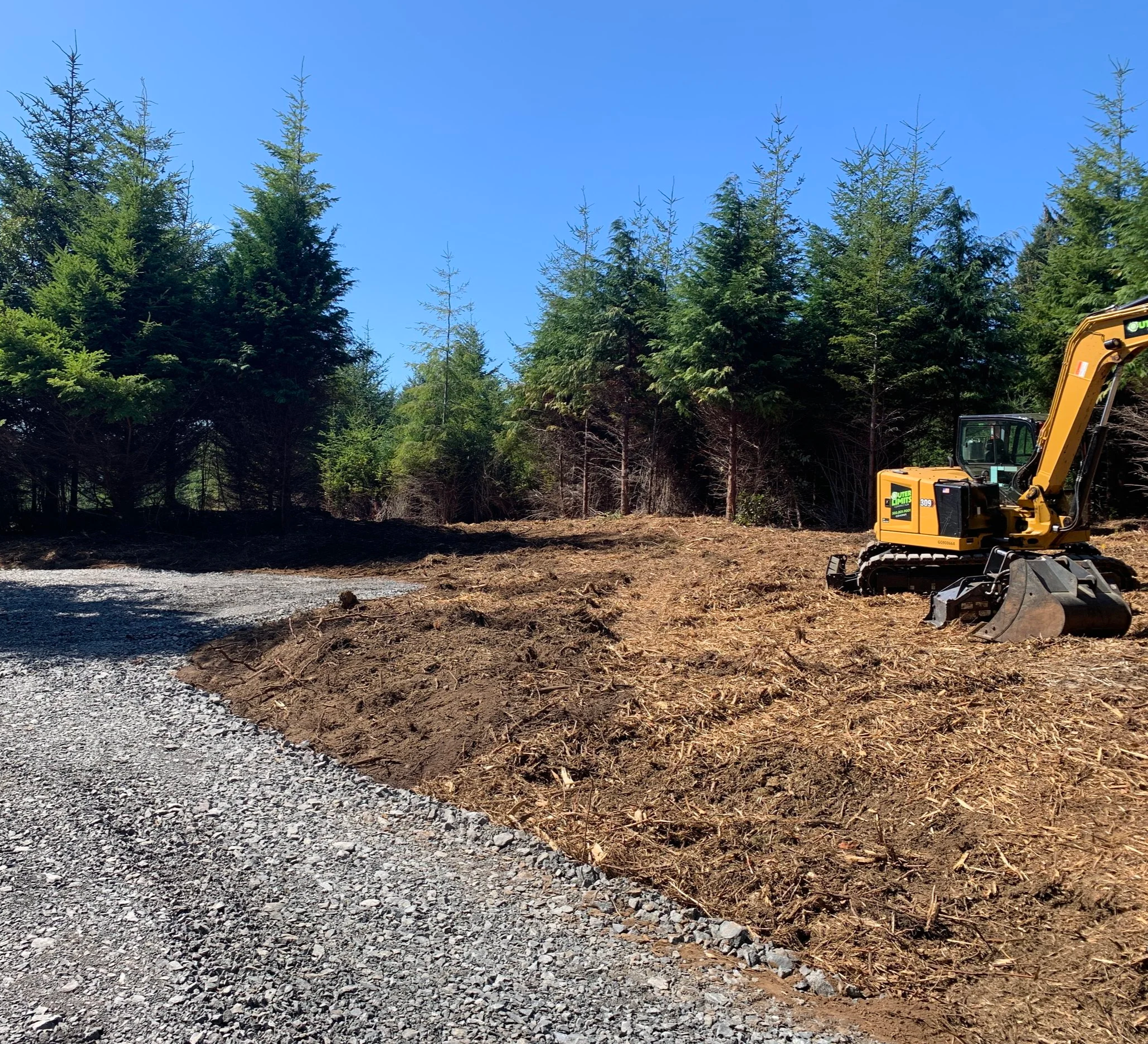 A small yellow excavator parked on a cleared dirt area beside a gravel path, with green pine trees in the background under a clear blue sky.