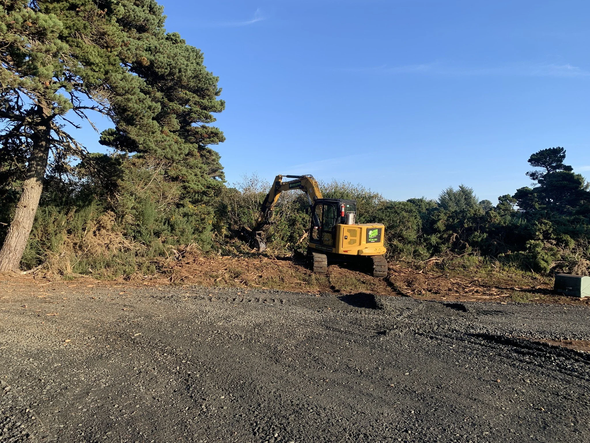 A small yellow excavator digging into a patch of dirt and shrubs near a gravel road under a clear blue sky.