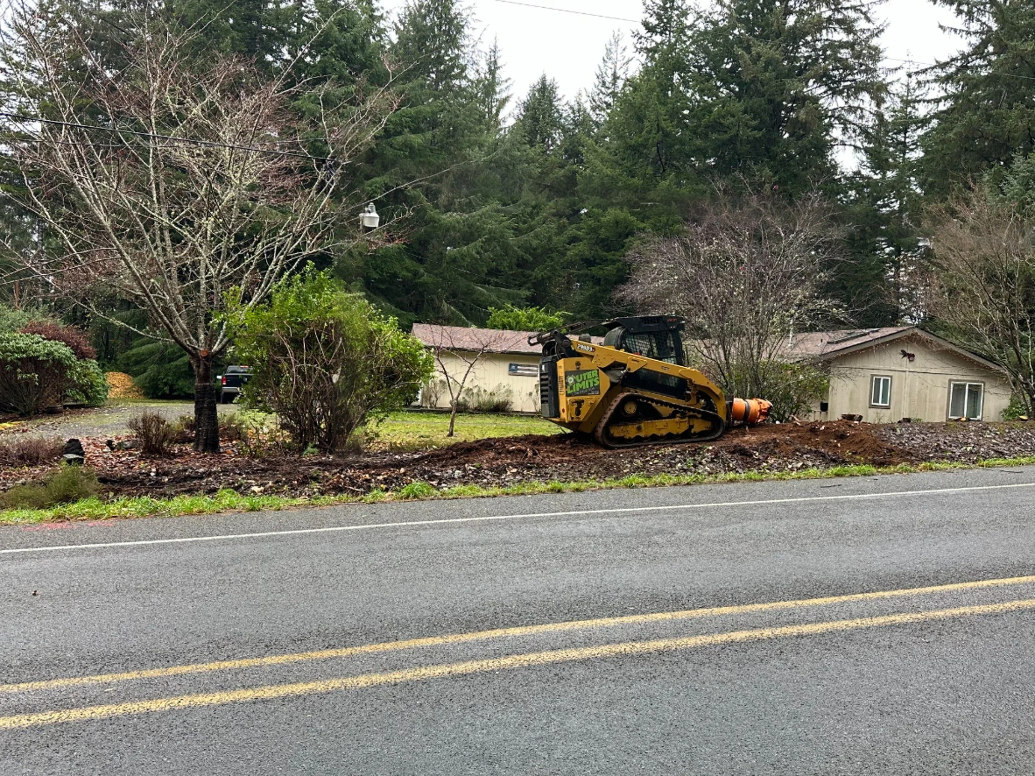 A construction vehicle, a yellow compact excavator with tracks, is parked on a dirt patch next to a road, with trees and houses in the background.