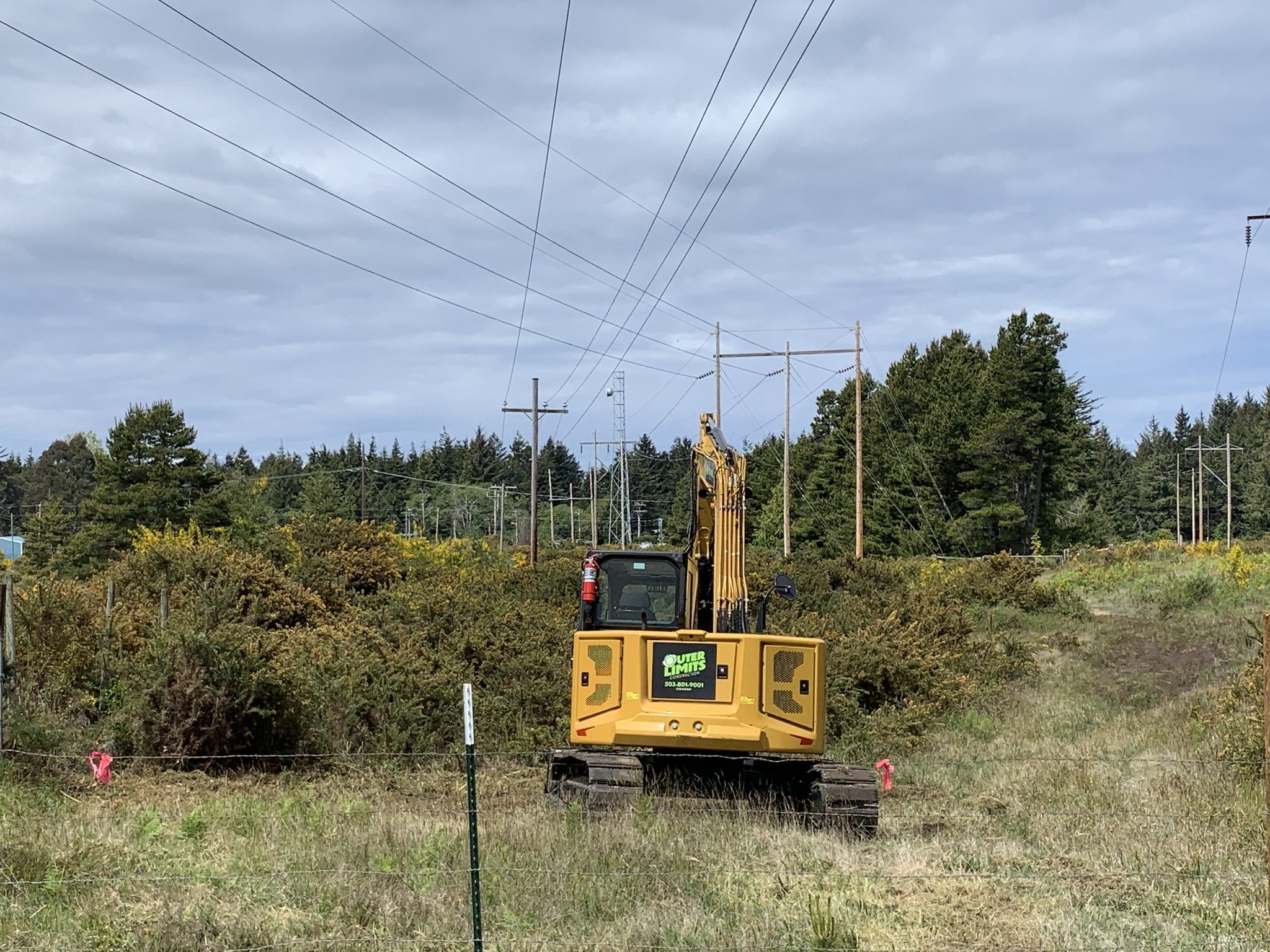 A small yellow excavator working on a grassy field near electrical power lines and trees under a cloudy sky.
