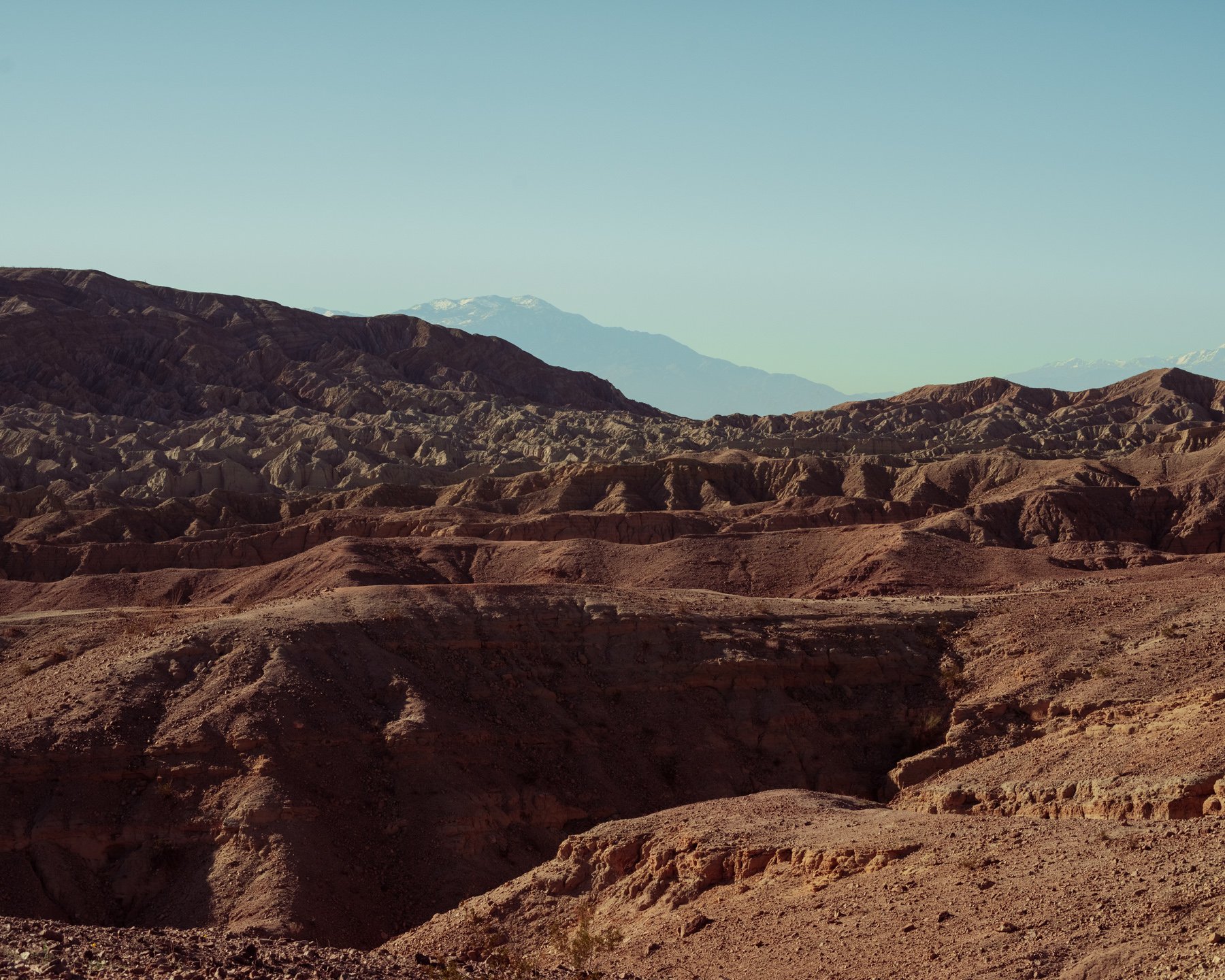 Desolate desert landscape with layered reddish and beige sedimentary hills, distant snow-capped mountains under a clear blue sky.